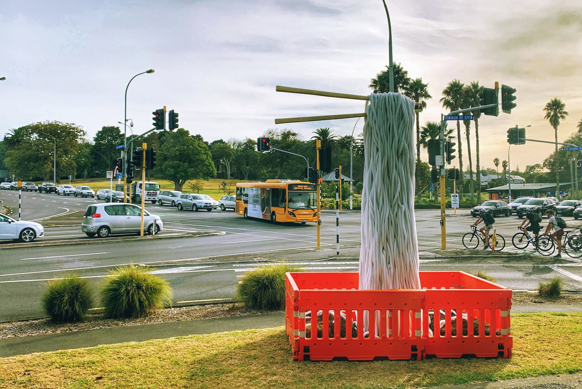 A view of the Balmoral-Dominion Roads intersection which includes part of a grassy area on a corner where orange roadwork barricades surround a tower of noodle-like objects being lifted by giant chopsticks held by an invisible hand. Traffic, including cars stopped at traffic lights, a bus and cyclists appear in the middle ground. 