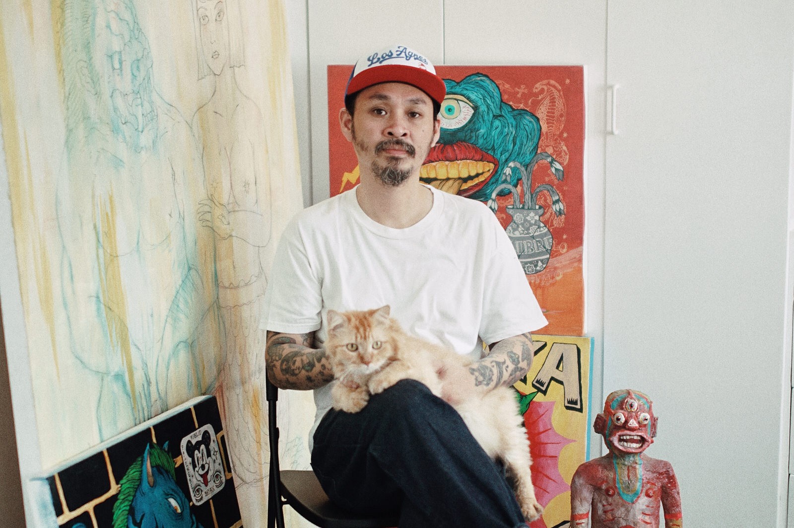 Man of Filipino descent with a cat on his lap sitting in front of various paintings