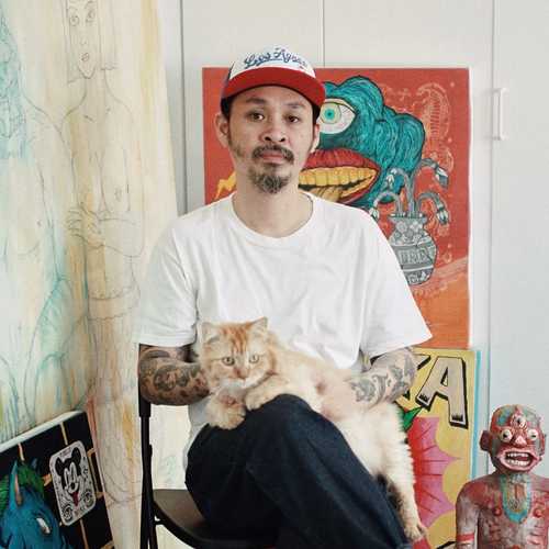 Man of Filipino descent with a cat on his lap sitting in front of various paintings