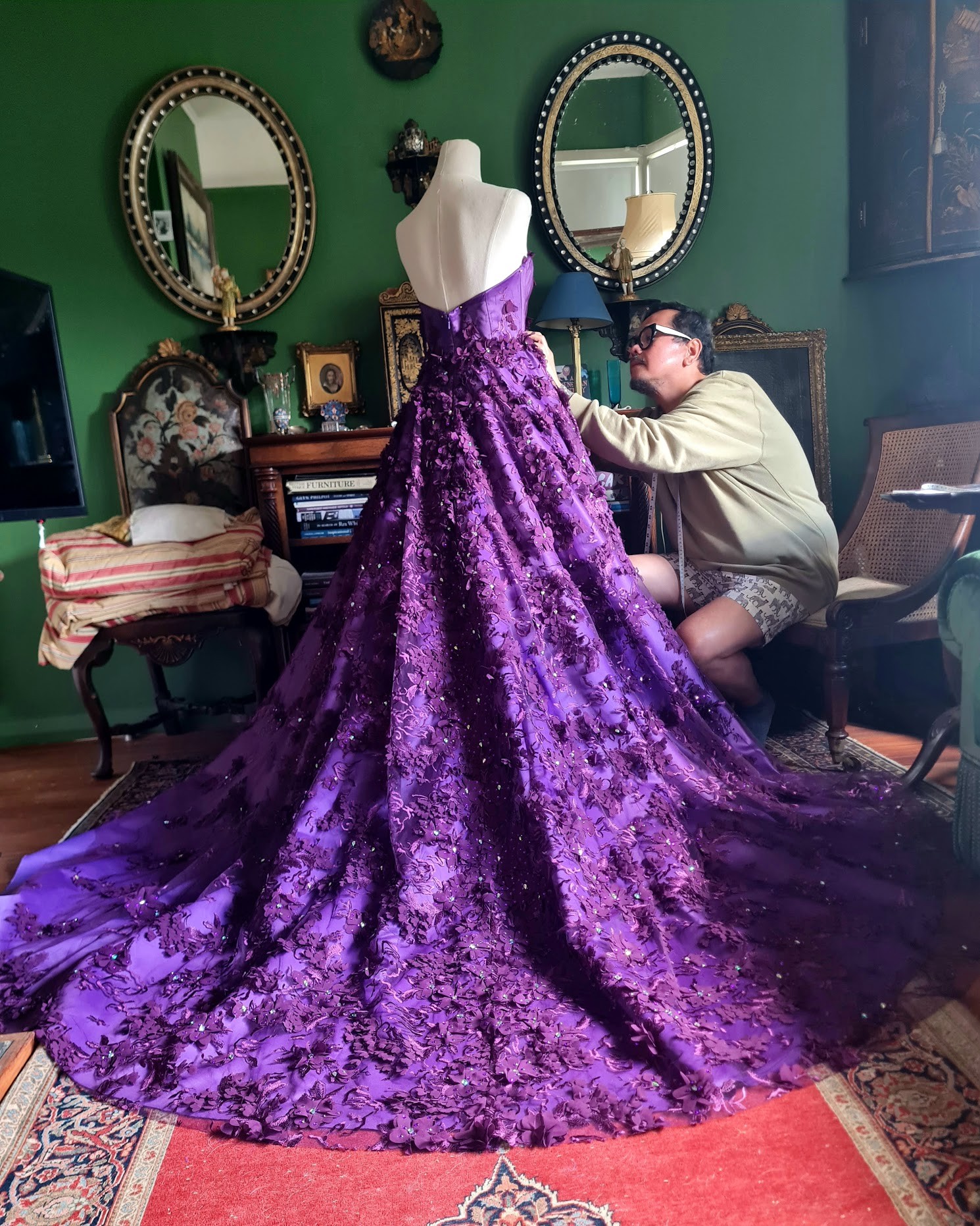 A man sitting on an antique chair is sewing a flowing purple gown with a long train