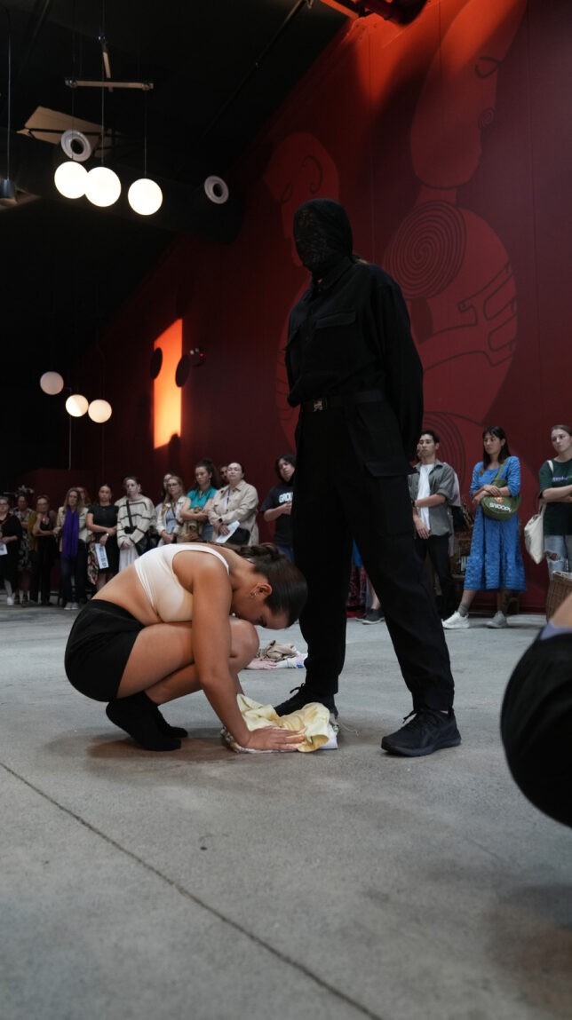 In a theatre foyer, a girl wearing cropped white top and black shorts crouching with her head and hands down. A masked officer in all black standing close to the girl, staring at her. Lots of audiences watching at the back.