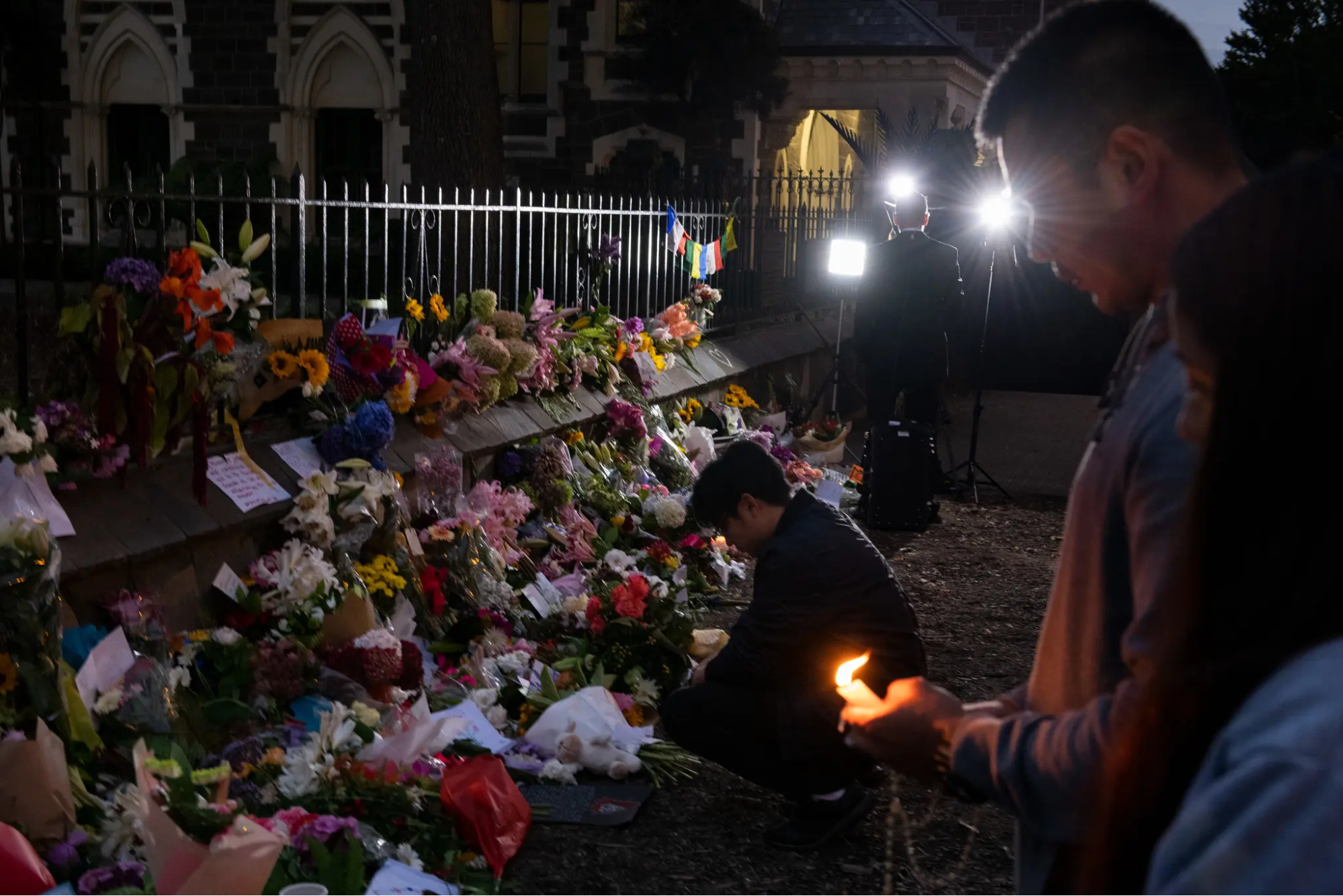 A person holds a lit candle while another lays flowers against a row of already-laid flowers