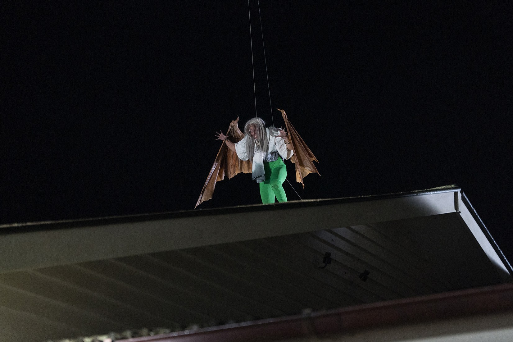 A horror movie set of a person with white hair and standing on a roof, wearing huge bat-like wings