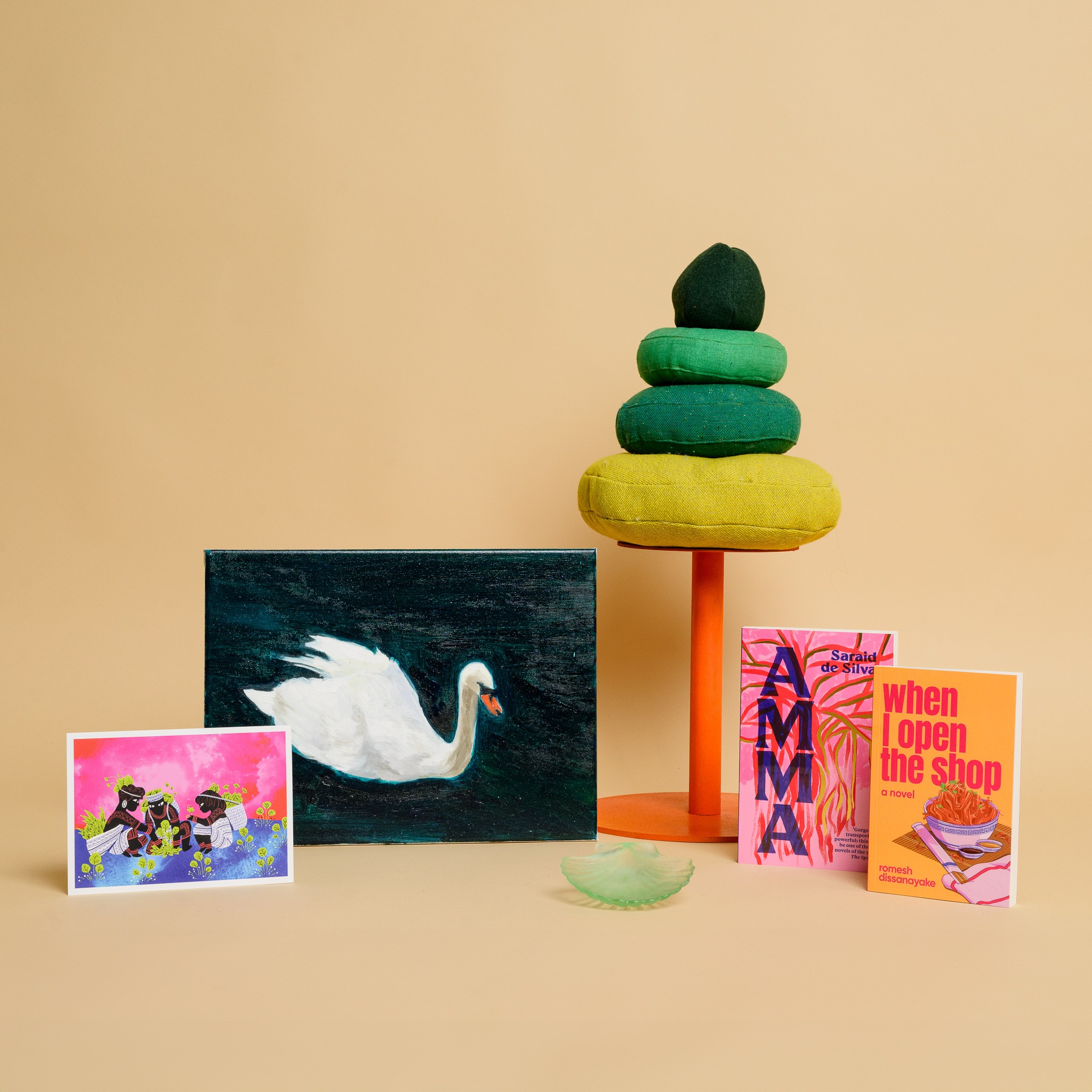 A painting of a white swan on emerald green water, a stack of three green cushions balanced on an orange plinth, two books, a green glass scallop and a print of three figures against a bright pink sky