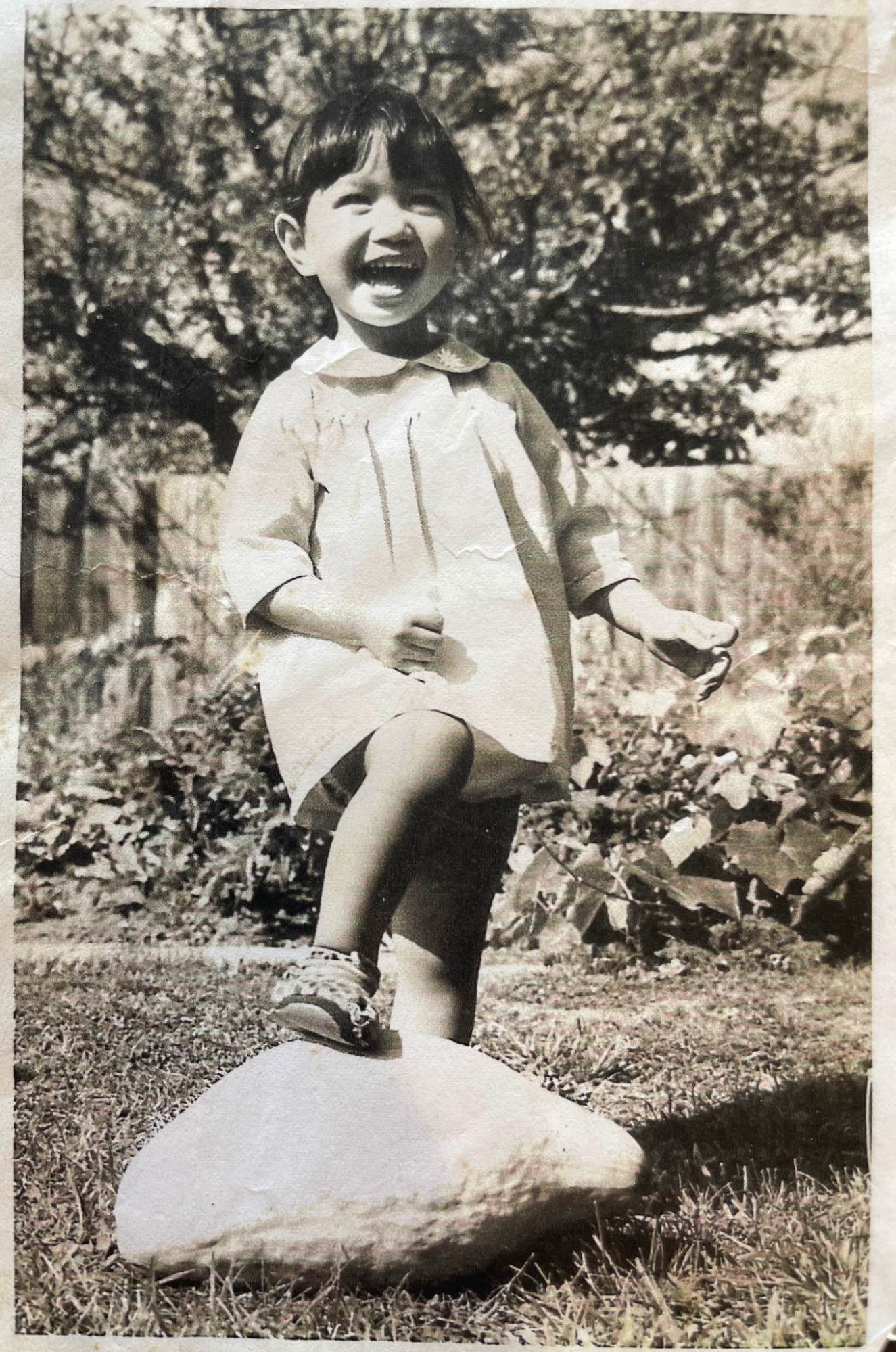 A sepia image of a laughing child in a garden, one foot on a rock in front of her