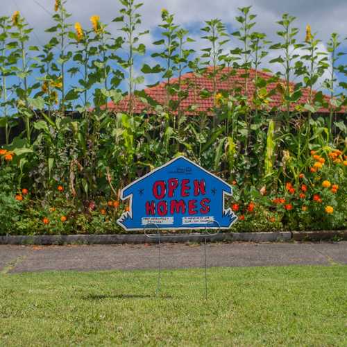 A blue sign that says 'OPEN HOMES' stands in front of a lush garden dotted with yellow flowers