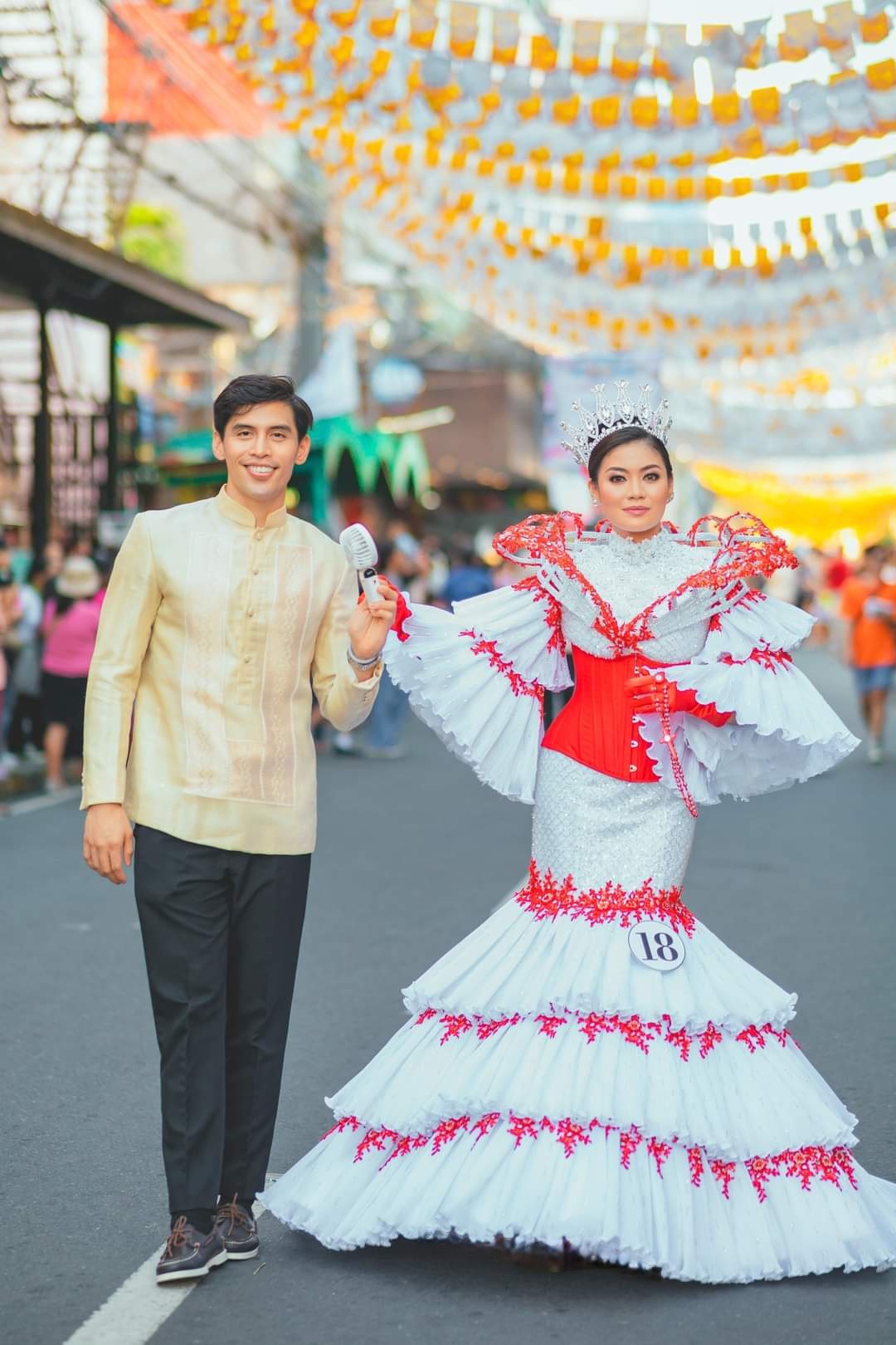 A woman wearing a silver crown and and an intricate white gown with red trimmings, her hand held by a man in a yellow jacket and black pants; behind them is a street with yellow buntings hanging from ceilings giving a festive scene