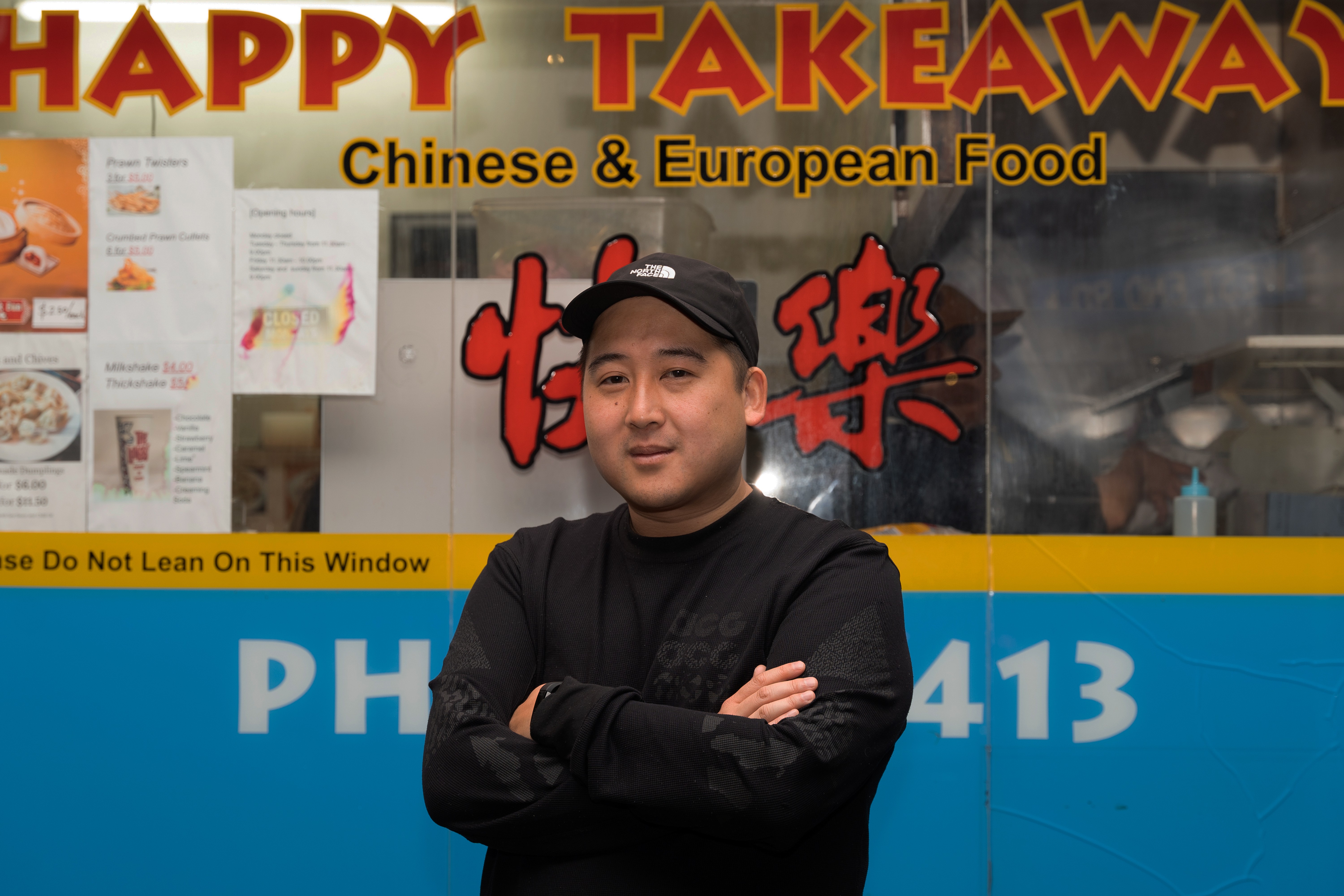 Lindsay Yee wearing a cap stands in front of a Chinese takeaway shop with his arms folded.