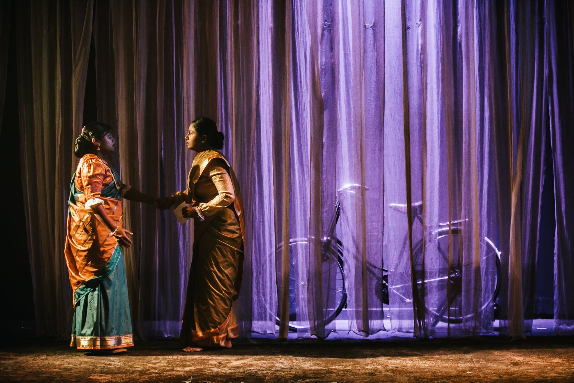 Two people in sarees on a stage in front of a curtain with a bicycle behind it