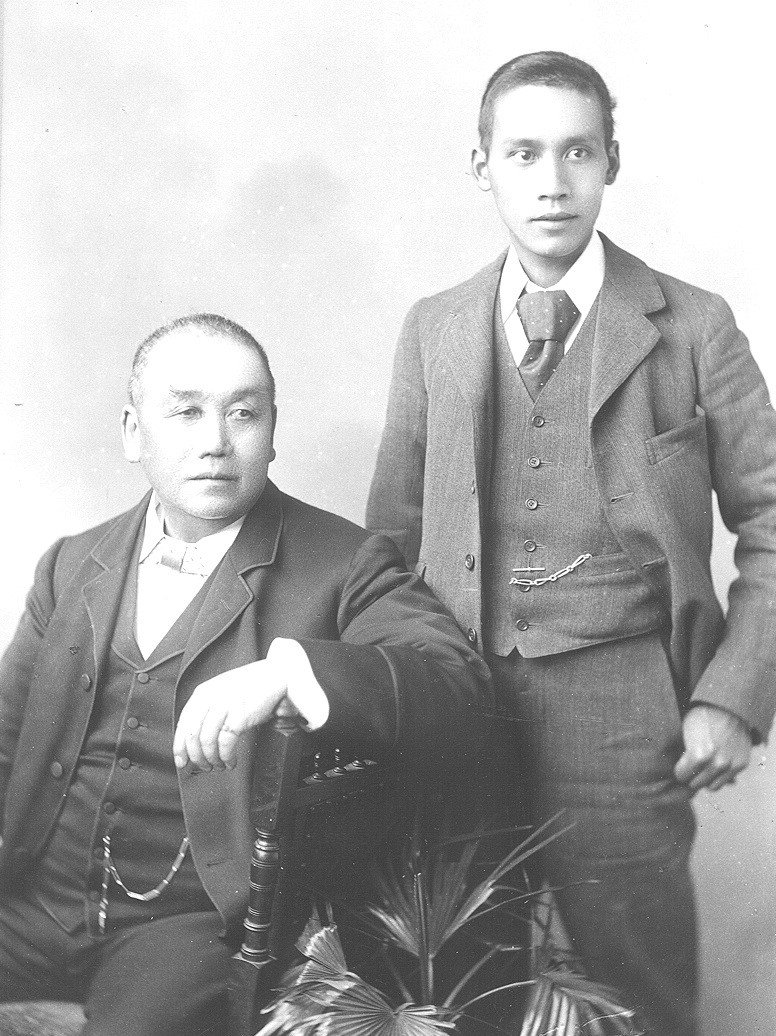 Two men in formal wear. One sits on a chair while the younger one stands behind.