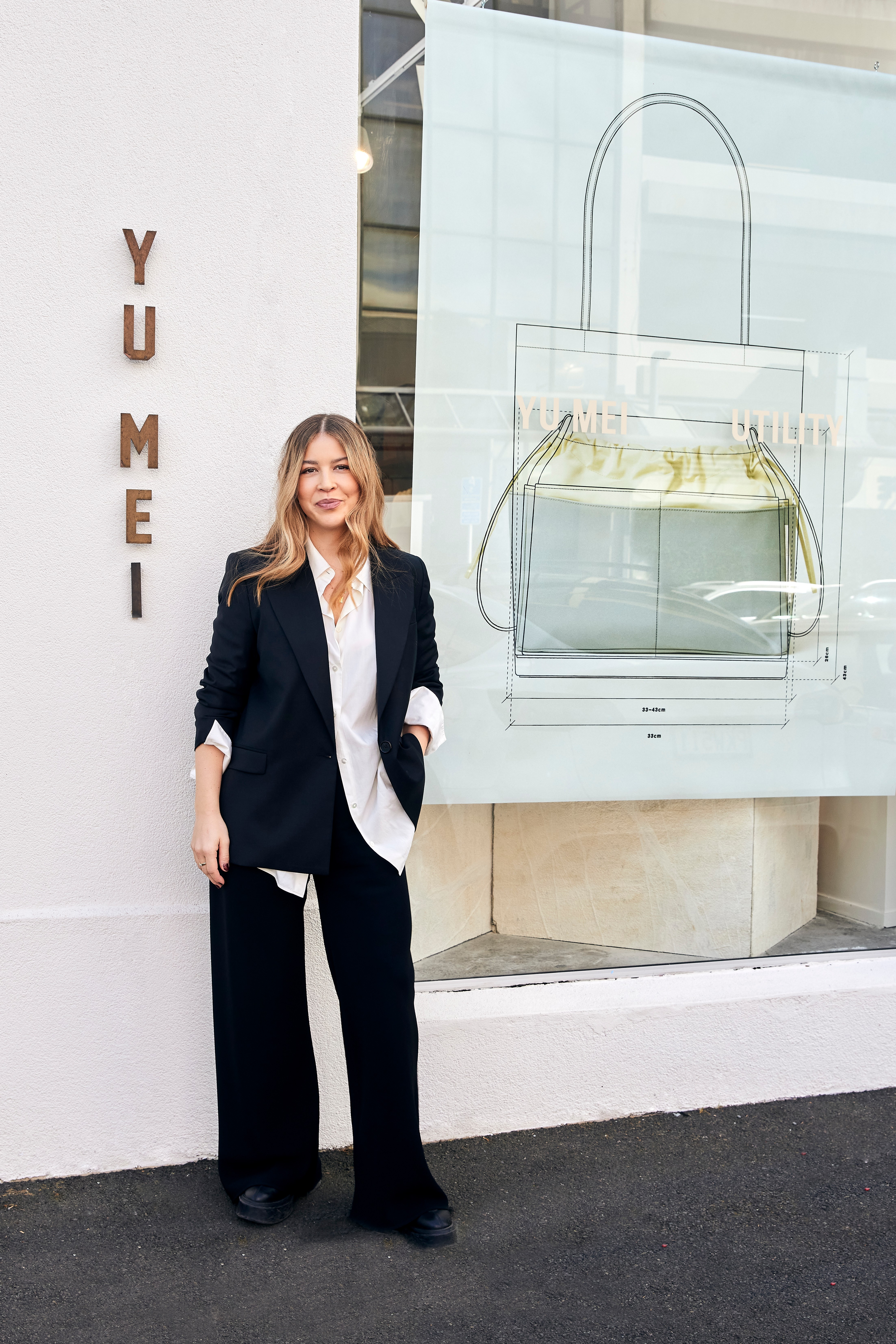 Jessie Wong, with blonde hair wearing a black and white suit, stands outside a Yu Mei handbag store.