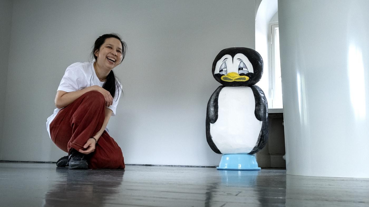 Woman sat on the floor laughing next to a sculpture of a cartoon-like penguin.