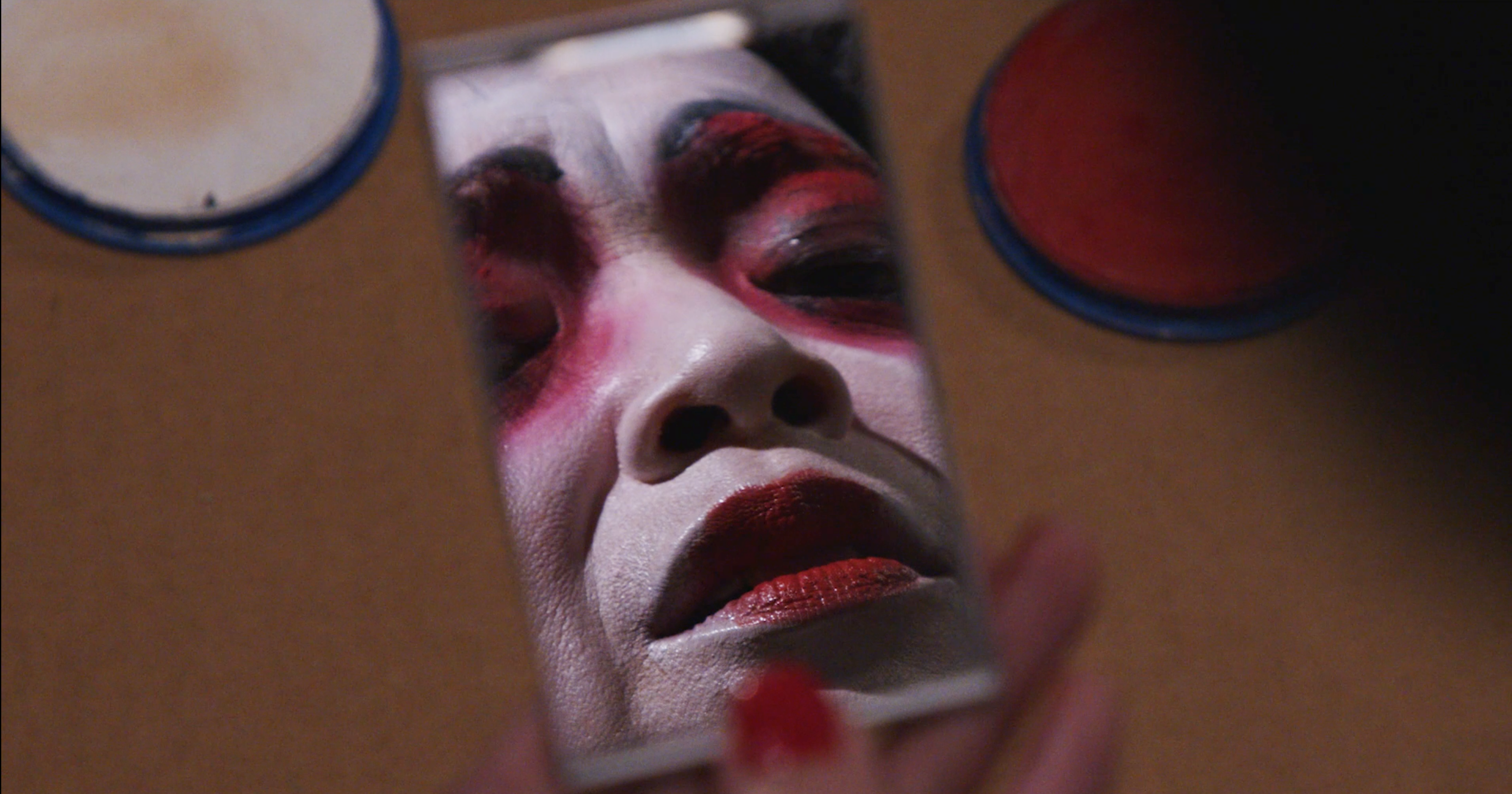 Image of a hand holding a small rectangular mirror. Reflected in the mirror is a woman whose face is painted white with red lipstick and bright red eyeshadow encircling her eyes.