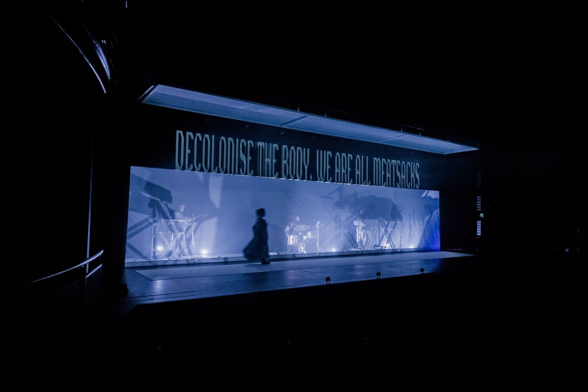 Atmospheric lighting on a dark stage with a projection that reads 'Decolonise the body we are all meat sacks'.