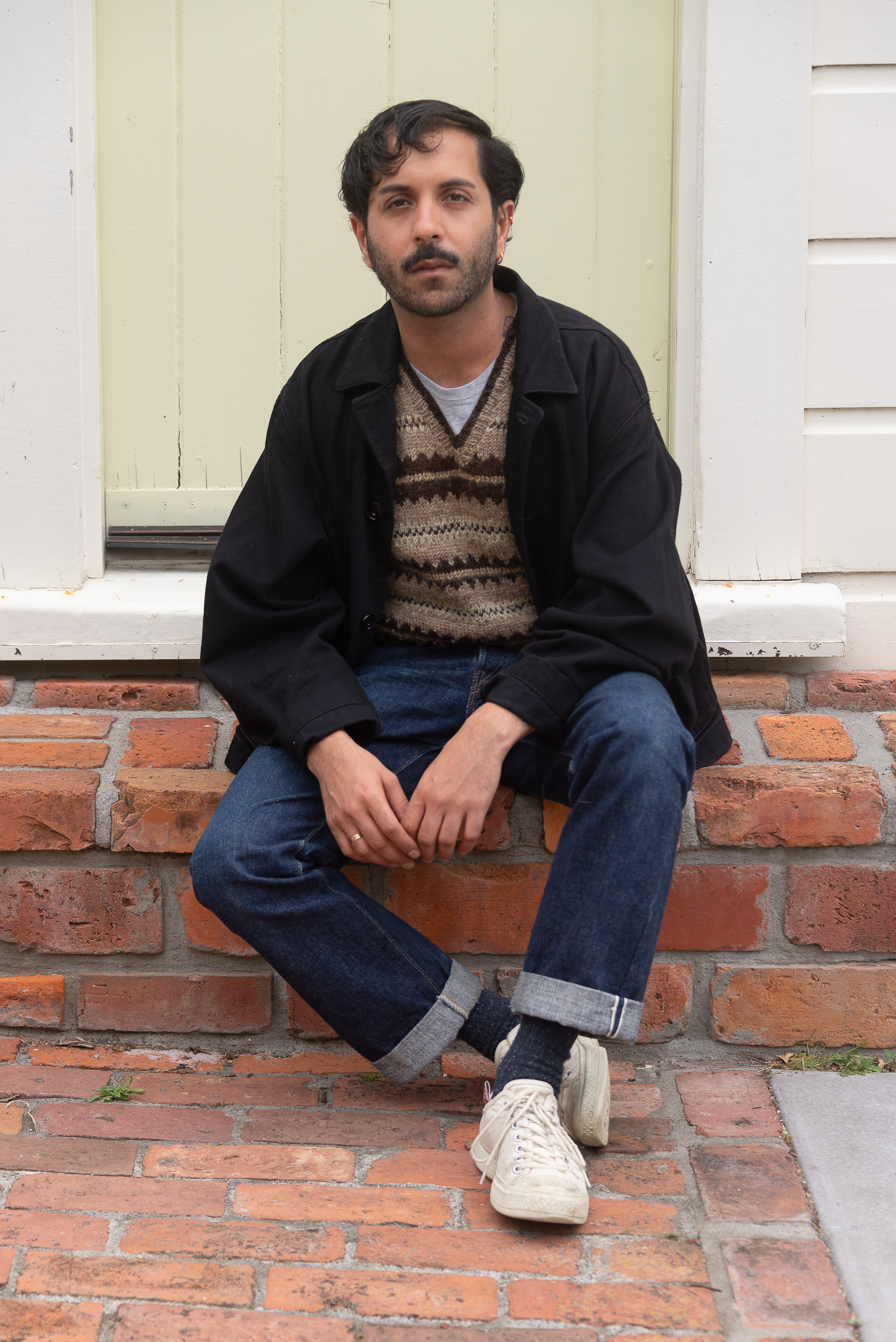 A person sitting on a brick door step in front of a pale yellow door. They have a beard and are wearing white shoes, blue jeans, a brown patterned jumper and a black jacket.