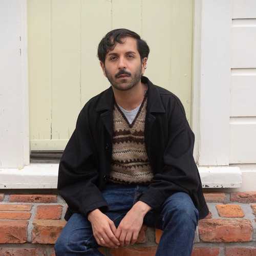 A person sitting on a brick door step in front of a pale yellow door. They have a beard and are wearing white shoes, blue jeans, a brown patterned jumper and a black jacket.