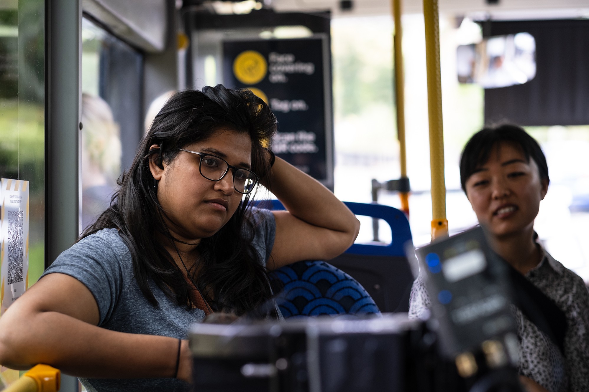 Two women on a bus, one wearing glasses and adjusting her hair while looking thoughtful, with camera equipment partially visible in the foreground.