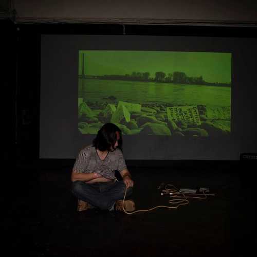 A man with shoulder-length hair sitting on the ground with a microphone recording his heartbeat in a dark space in front of a projection of his video work