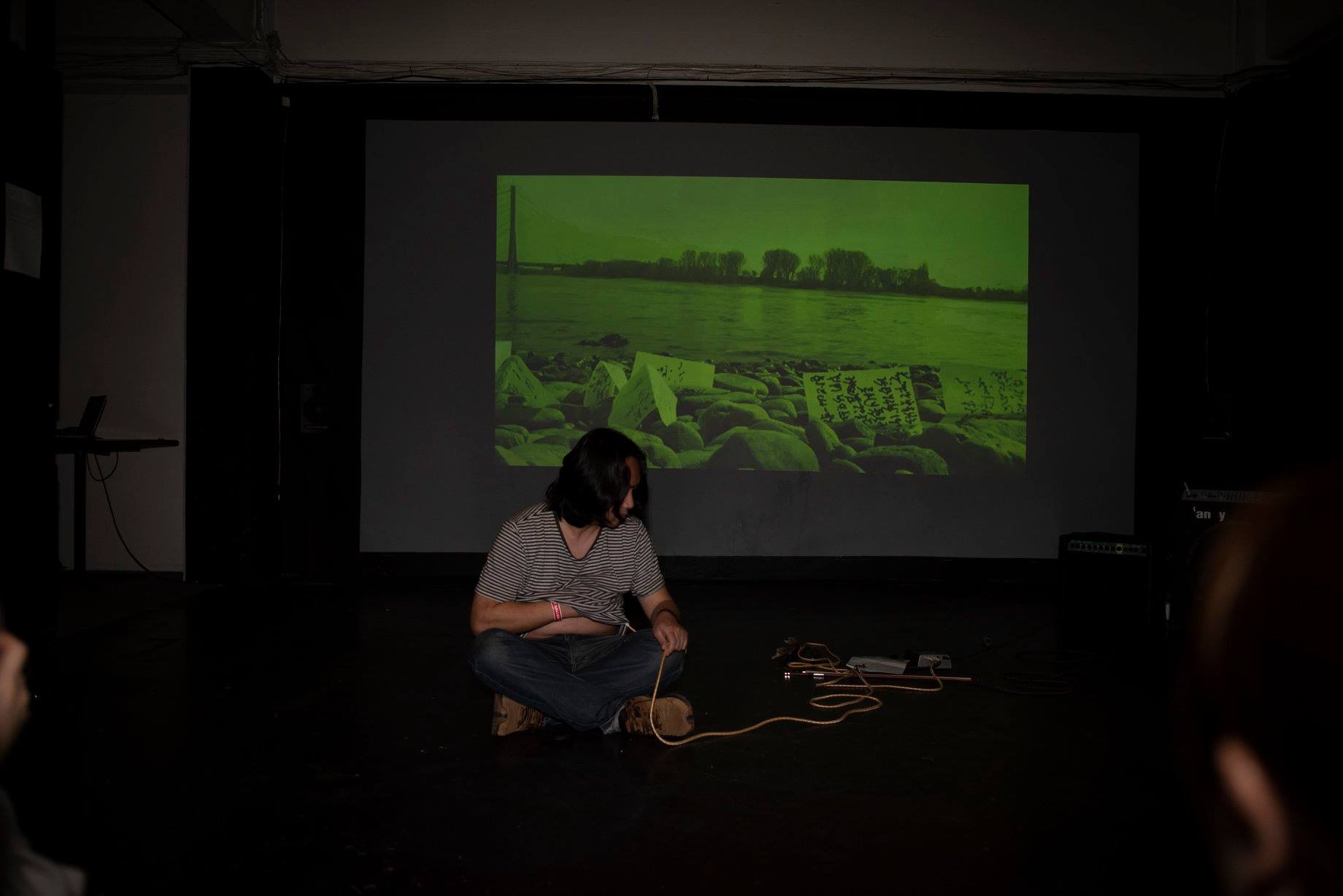 A man with shoulder-length hair sitting on the ground with a microphone recording his heartbeat in a dark space in front of a projection of his video work