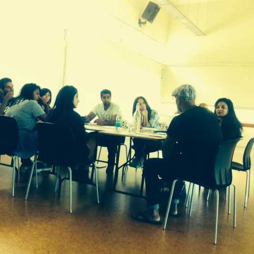 A group of about 10 South Asian writers and actors sit around a table, talking and looking at a script.