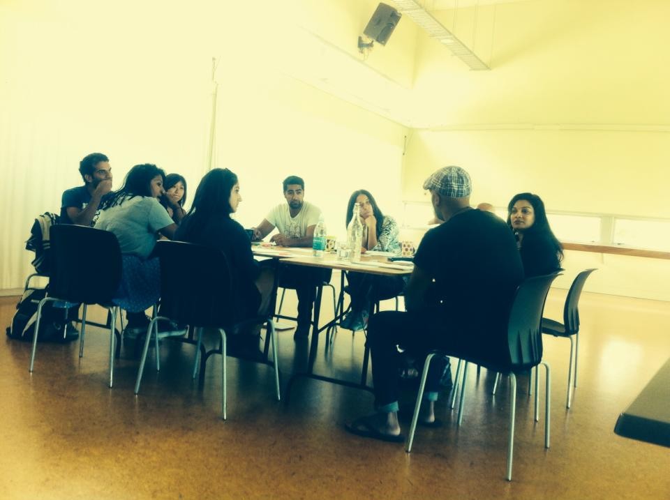 A group of about 10 South Asian writers and actors sit around a table, talking and looking at a script.