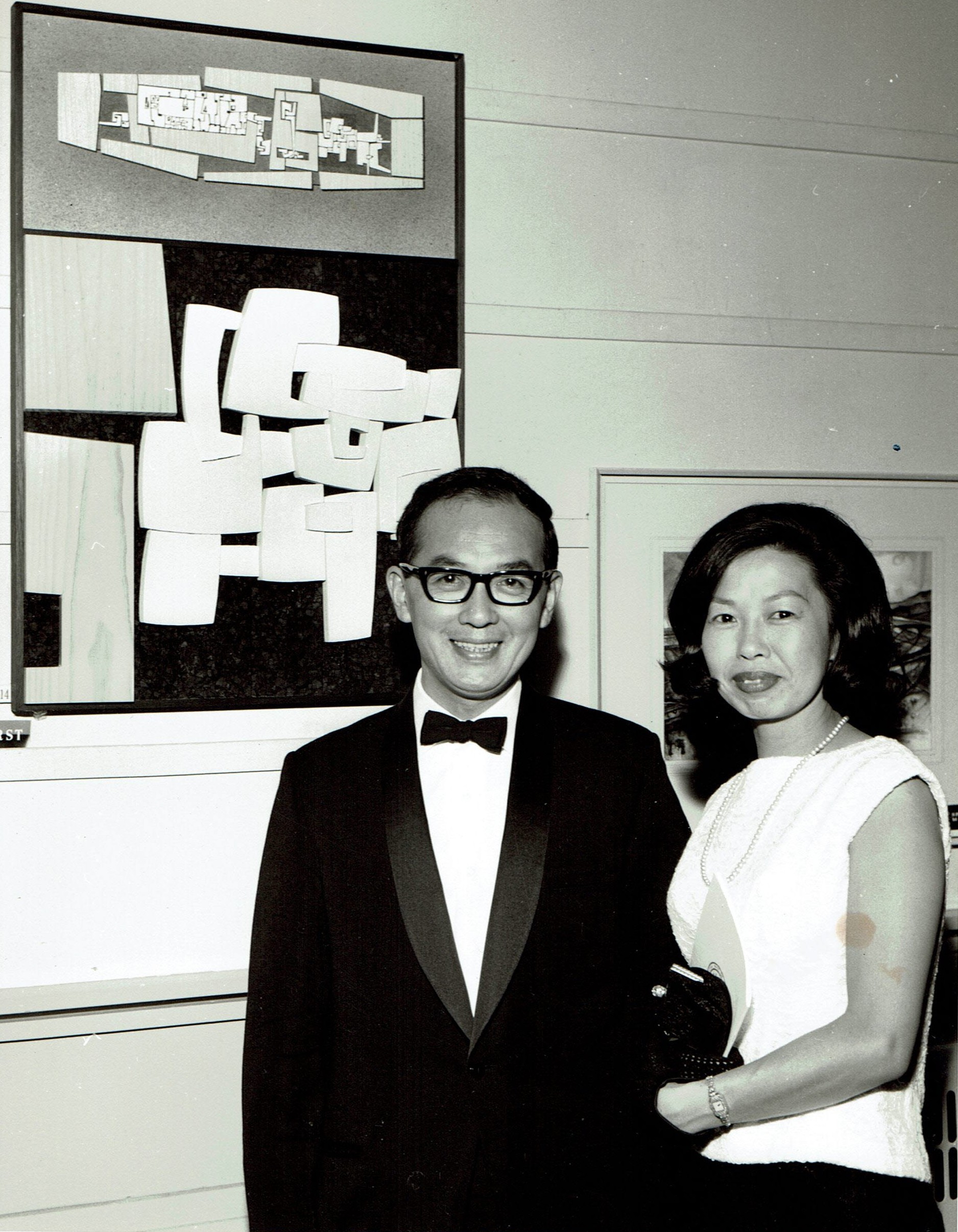 A smiling couple dressed formally stand arm-in-arm in front of an abstract painting