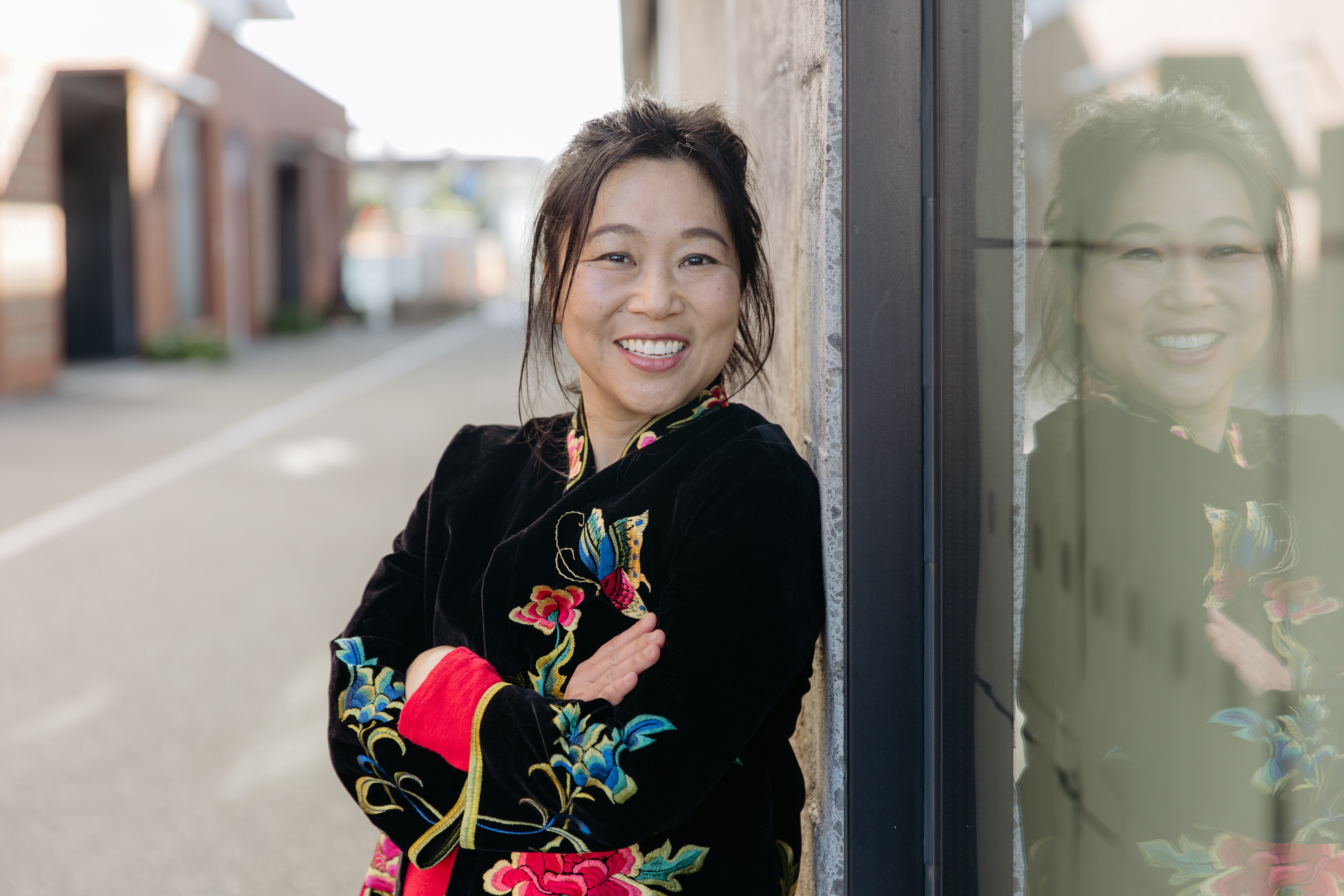 A woman leaning against a shop-front on a street, wearing a black velvet jacket embroidered with colourful flowers, smiling.