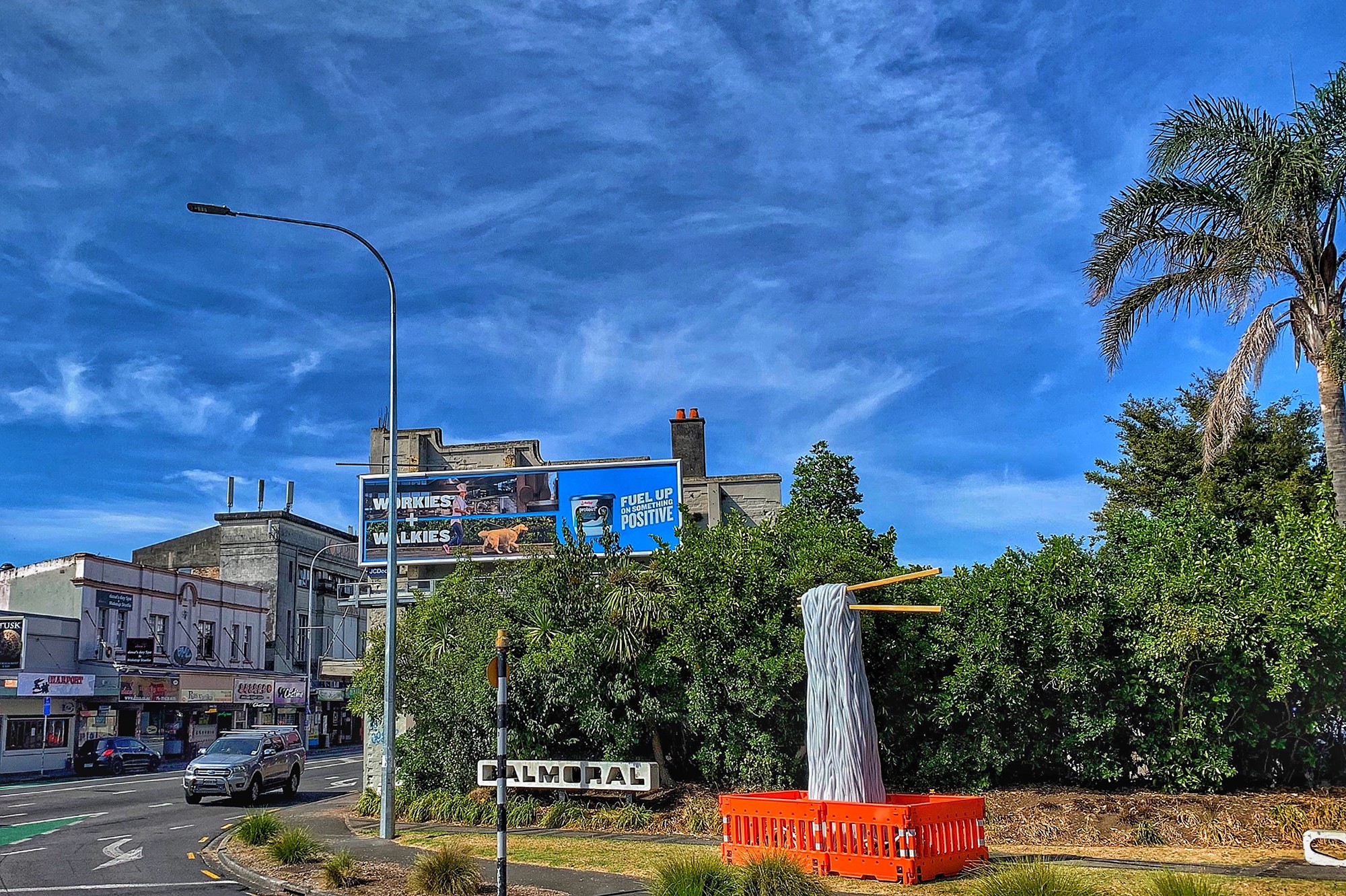 A grassy area with trees and a sign reading 'Balmoral' on the corner of a intersection where orange roadwork barricades surrounding a tower of noodle-like objects being lifted by giant chopsticks held by an invisible hand. A large billboard is on the side of a building in the middle ground, and a shop lined street, Dominion Road, is visible on the left side of the image with a car coming around the corner.