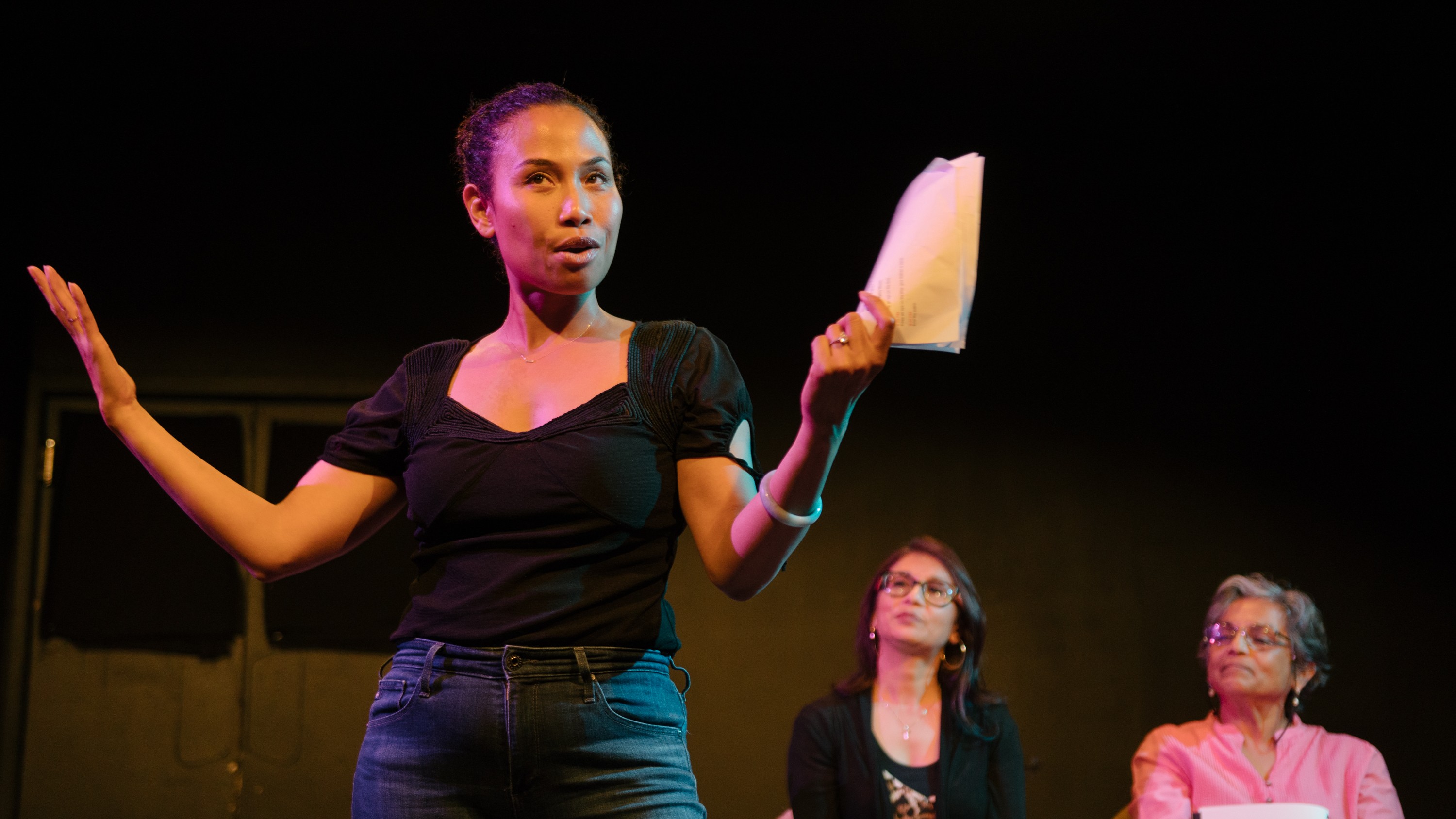 A woman holding a script stands gesturing to the audience while two other people sit in the background observing