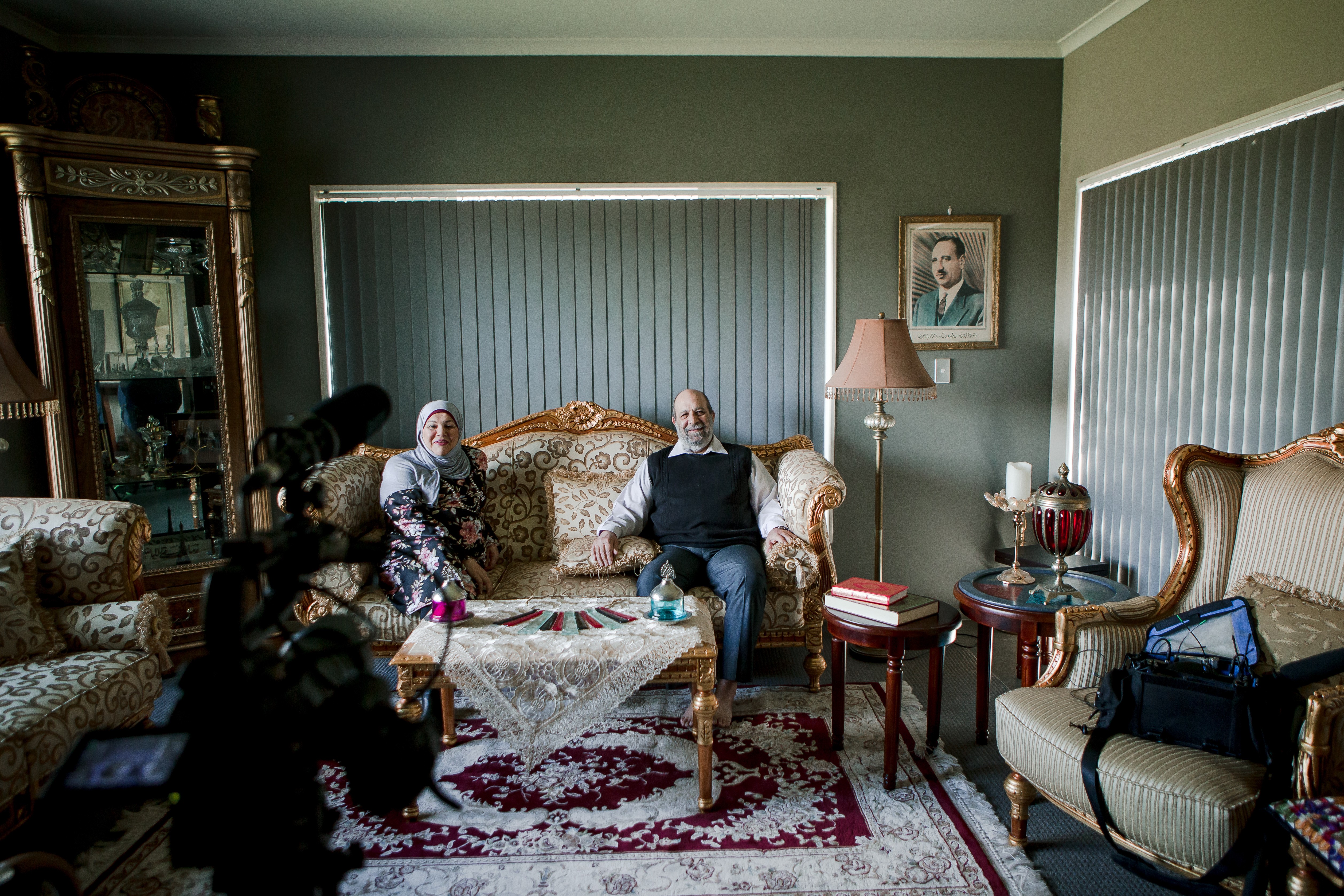 A woman in a headscarf and a man sit on a couch in a living room, with a big Persian rug. Sound and filming equipment faces them.