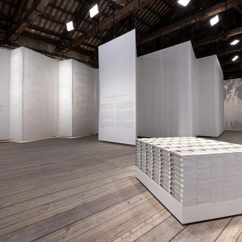A room in which large scrolls of white paper hang from the roof to create a wall. In the centre of the room, a brick of books creates a rectangular form on the ground.