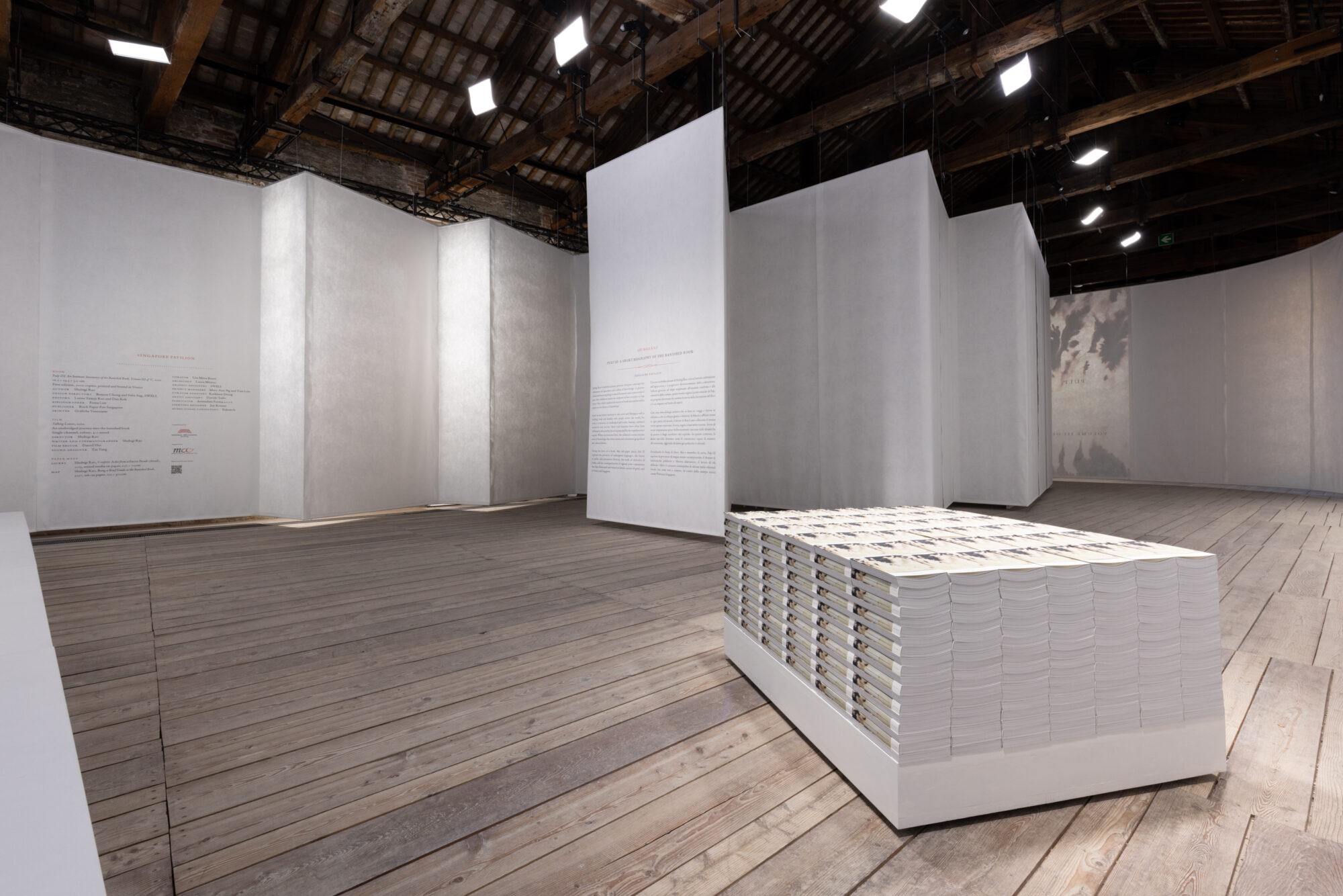 A room in which large scrolls of white paper hang from the roof to create a wall. In the centre of the room, a brick of books creates a rectangular form on the ground.