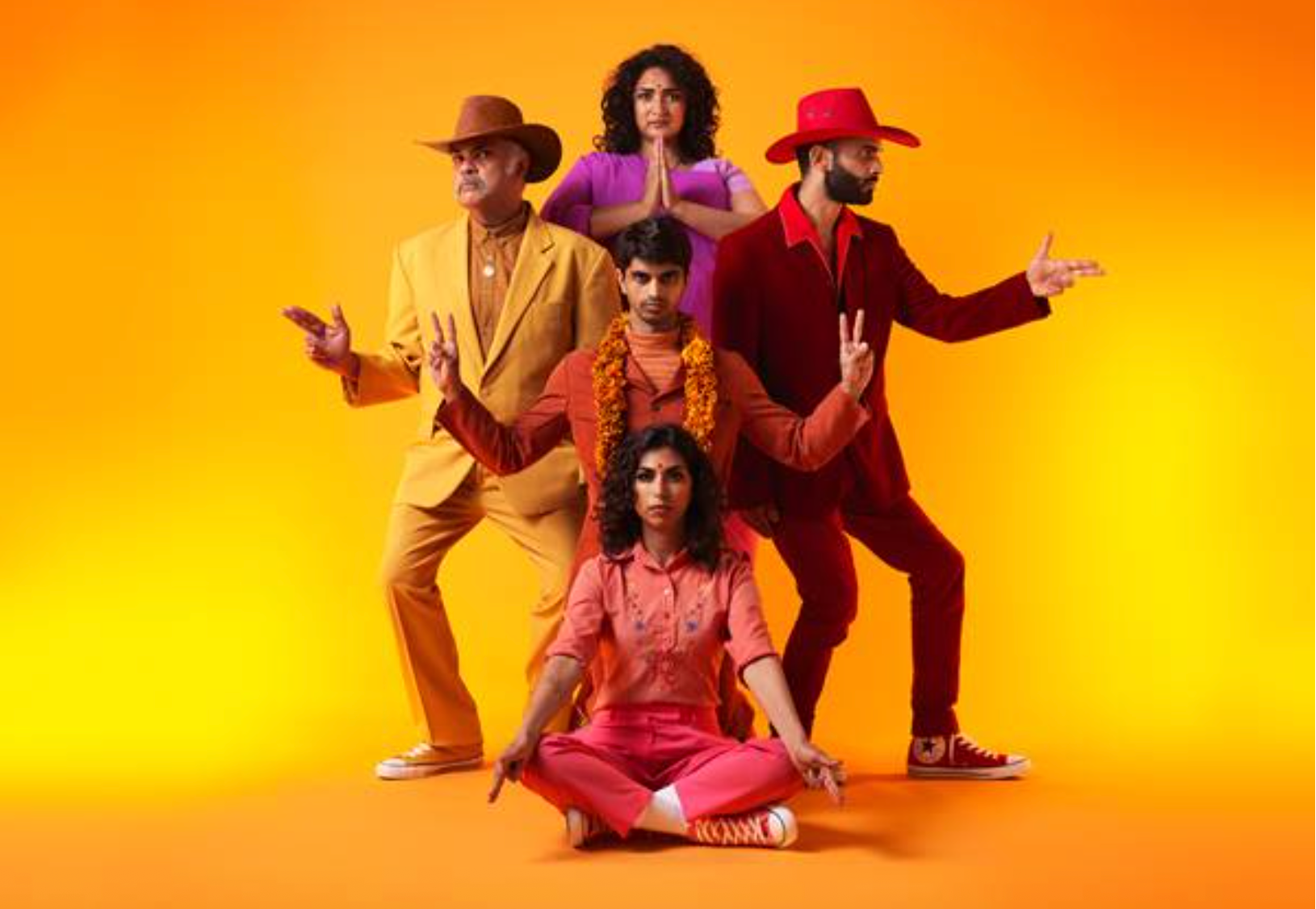 Five South Asian actors in warm-toned Desi Cowboy outfits pose direct to camera, throwing peace signs, prayer signs and finger guns against a bright orange background.