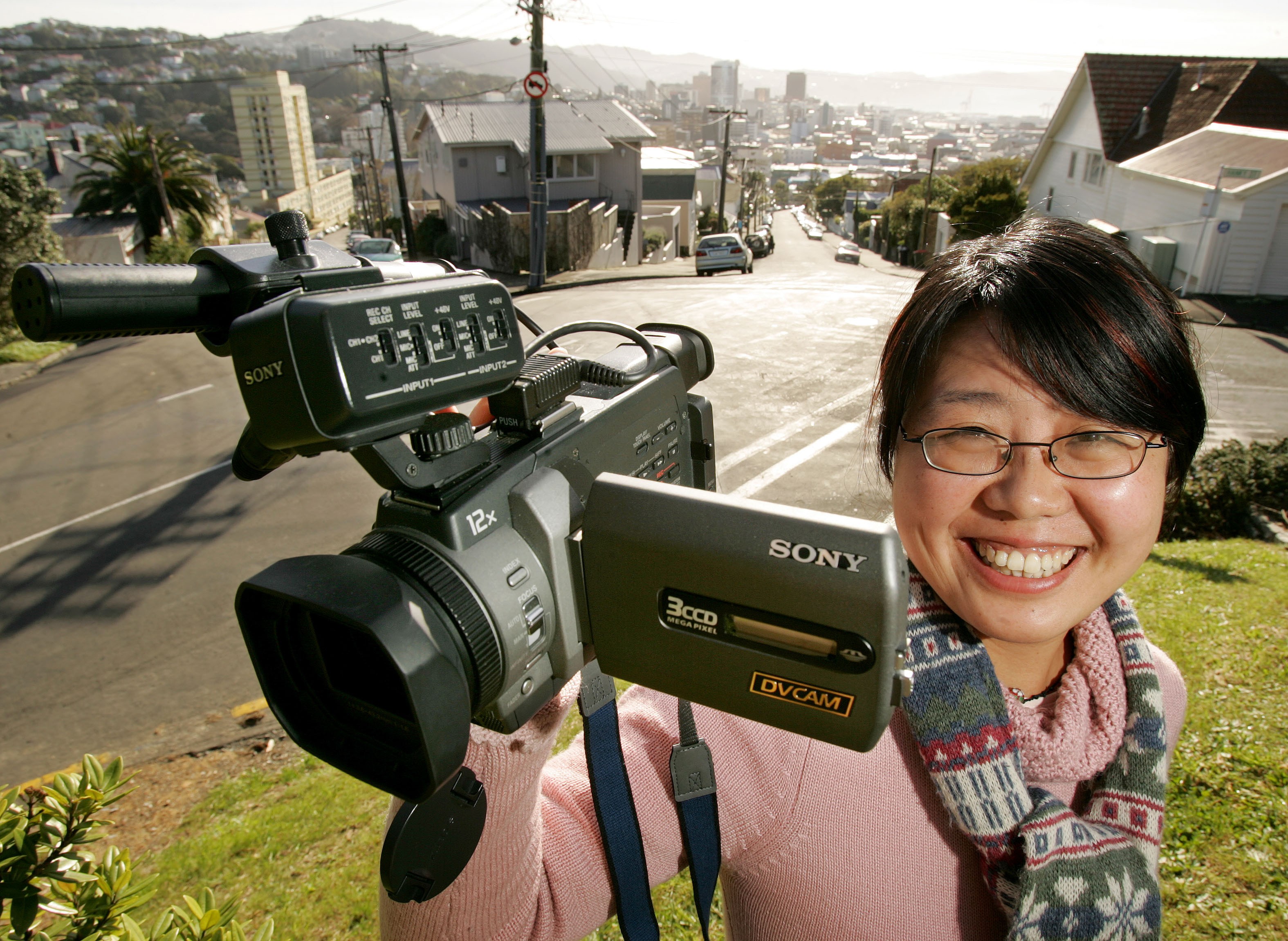 A woman wearing glasses and a pink sweater holds a Sony digital video camera in her right hand, in front of a residential street. 