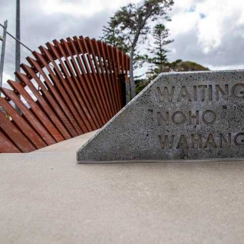 A concrete plaque that says 'Waiting Noho Wahangū Ana' with a curved series of narrow steel panels behind