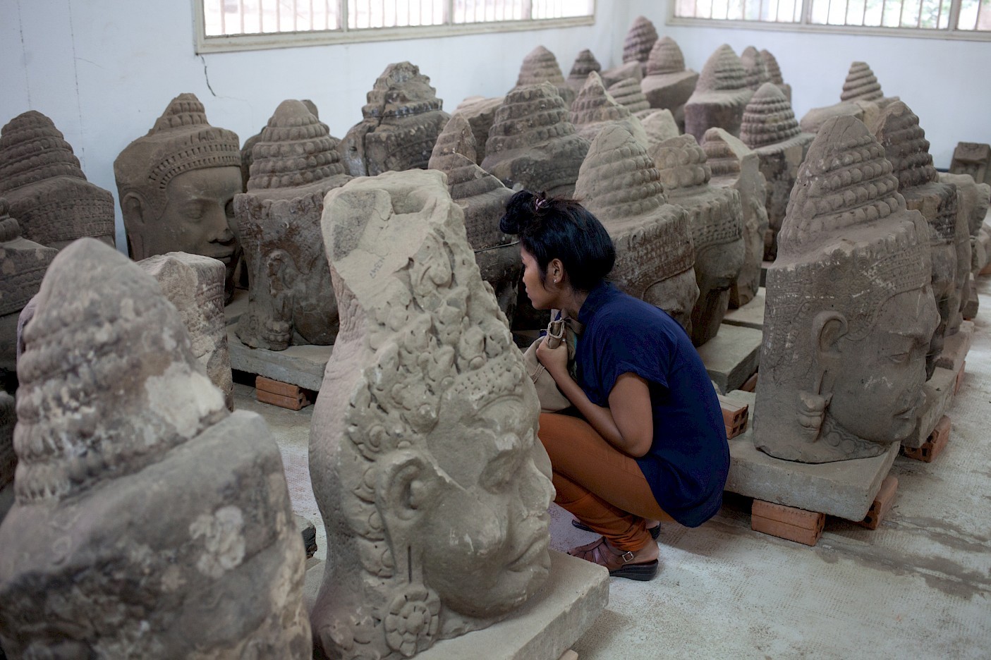 A woman crouches among large stone heads in a small room.