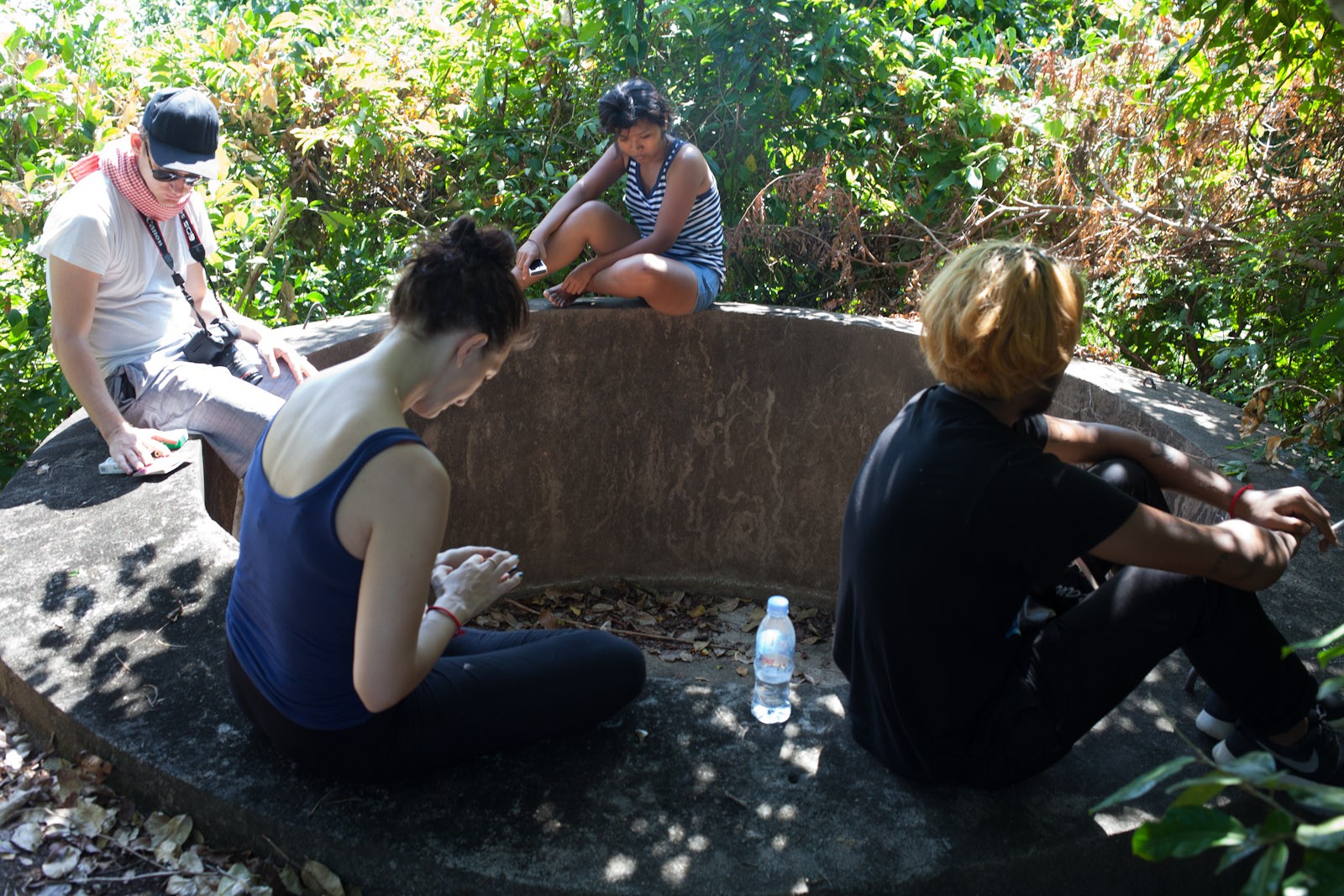 A group sit around a well-like structure amongst overgrown plants.