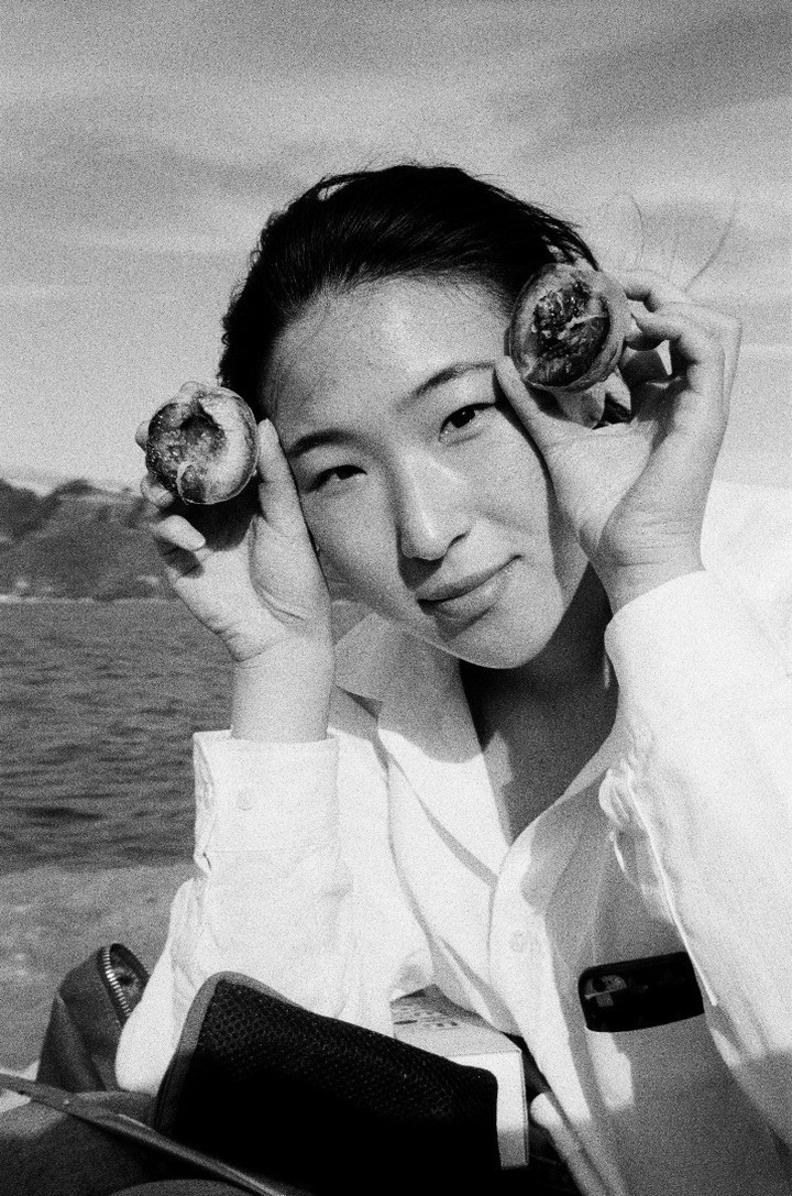 Black and white photo of Emerita Baik in front of the ocean holding up two halves of a nectarine.