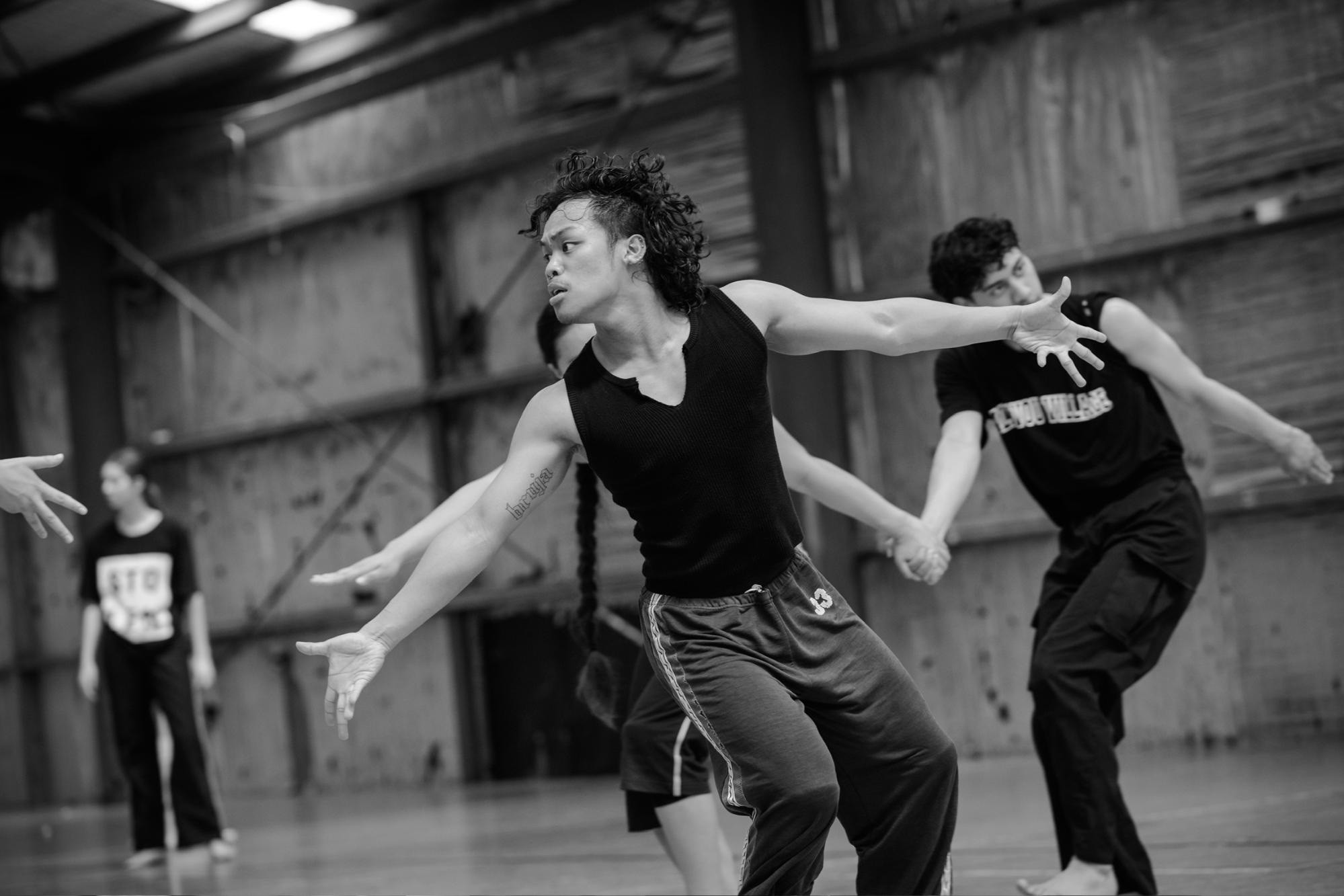 Black and white action photo of a Filipino-person in the foreground dancing with arms outstretched along with other dancers in the background