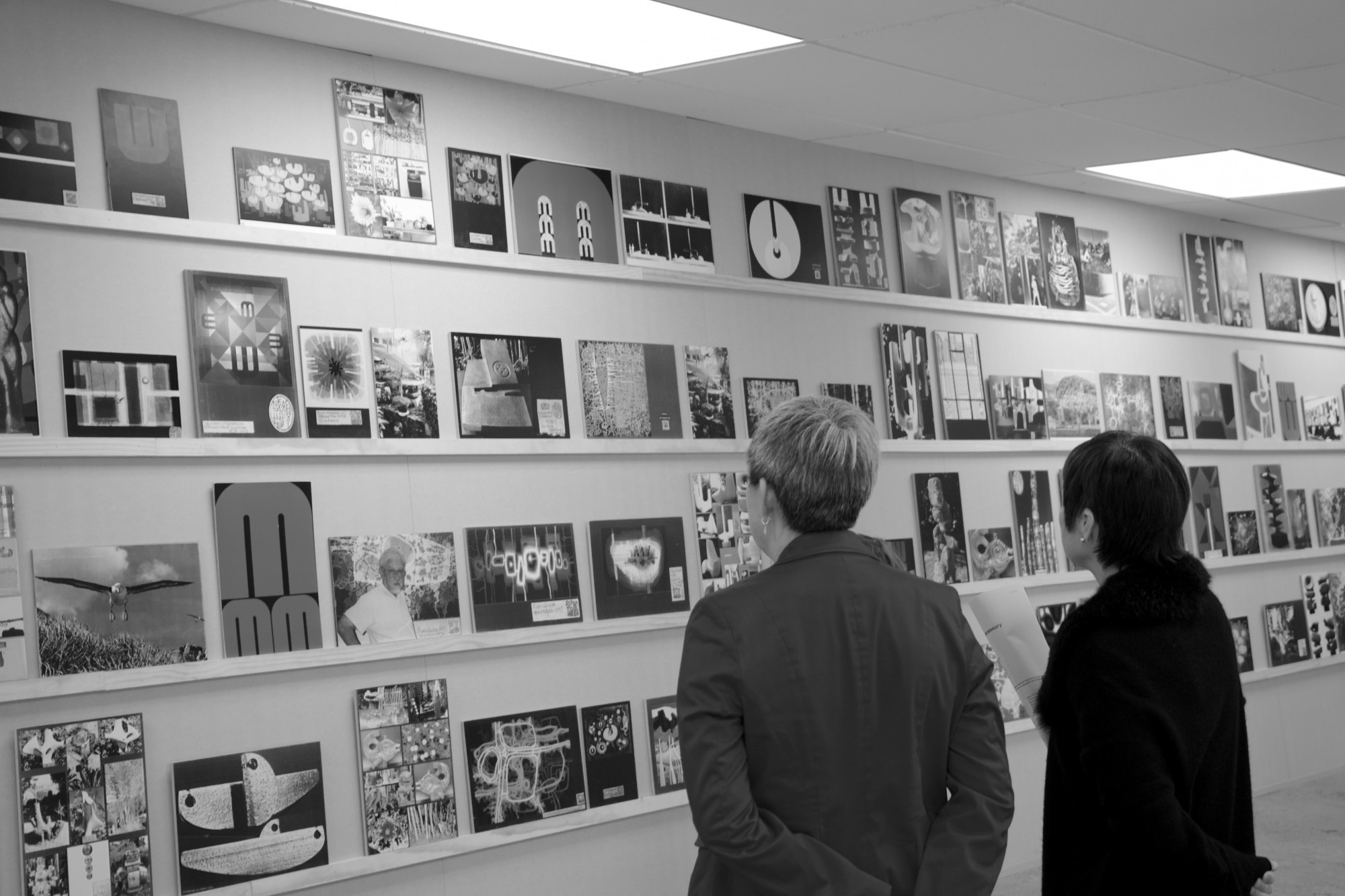 Two women with their back to us stand looking at shelves with photos of works by Guy Ngan laid along them.