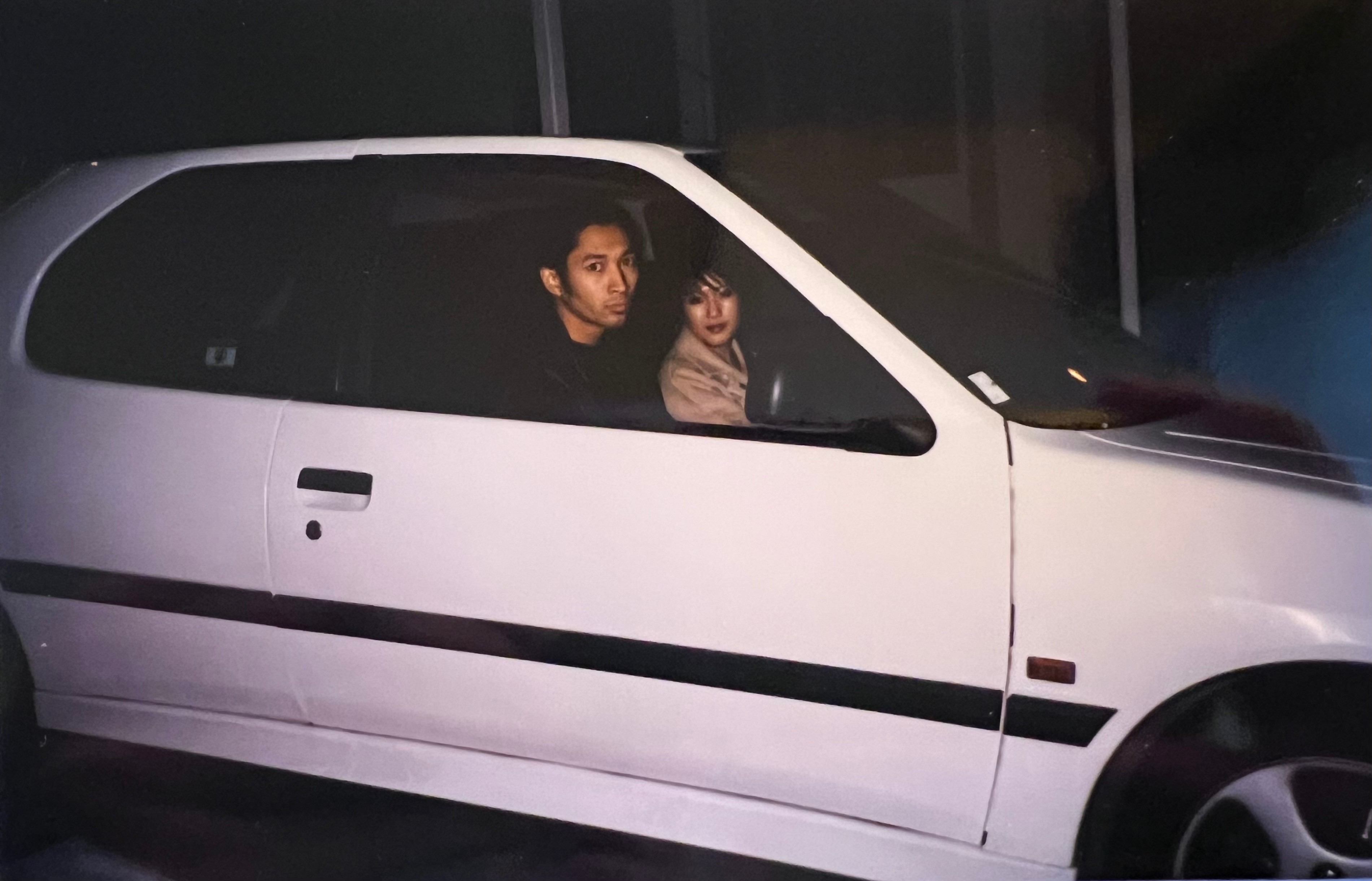 A man and a woman sit inside a white hatchback car.