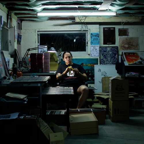 A person in a dimly lit artist studio creating work. Various pieces of art and posters are on the walls.