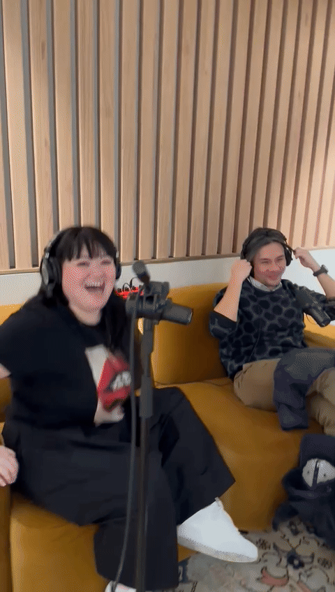 A man and a woman sitting on a mustard couch with headphones on, laughing