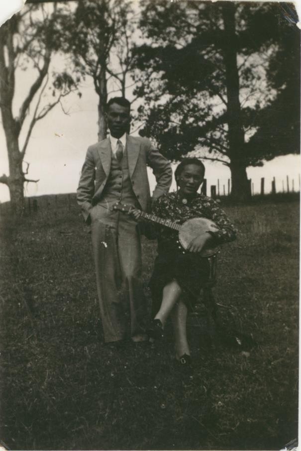 A man stands on a hillside next to a woman who is sitting playing the banjo