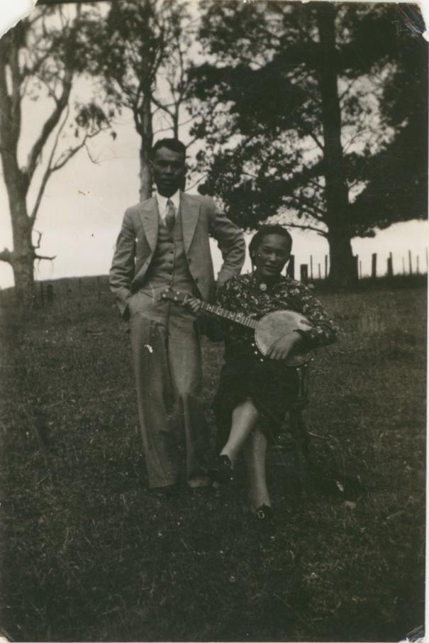 A black and white photo of a man and woman on a hill. The man is standing and the woman is sitting playing a banjo