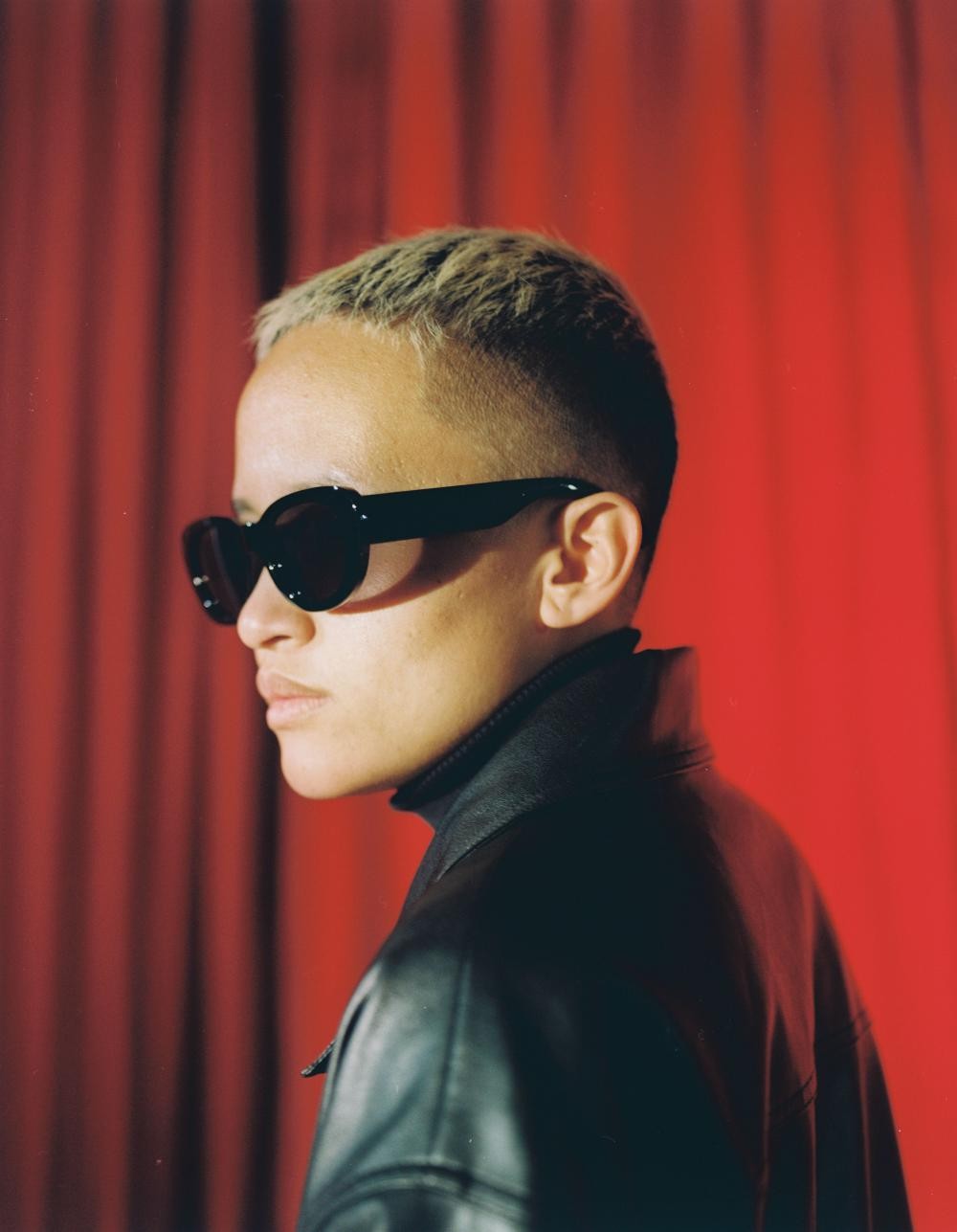 Person of Filipino-descent with close cropped hair, dark glasses and wearing a black leather jacket posing in front of red curtains