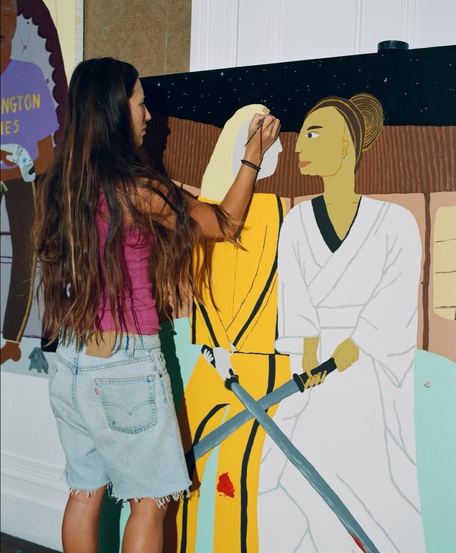 A person with long dark hair stands in front of a large canvas, painting a picture of two figures based on characters from the movie Kill Bill