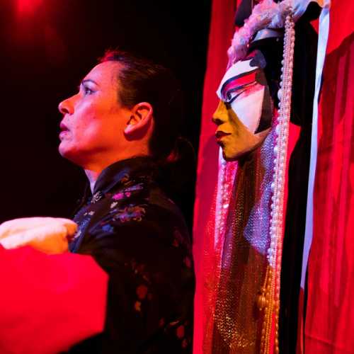 A woman stands on stage performing a play. Behind her hangs red set dressing and a Chinese opera mask.