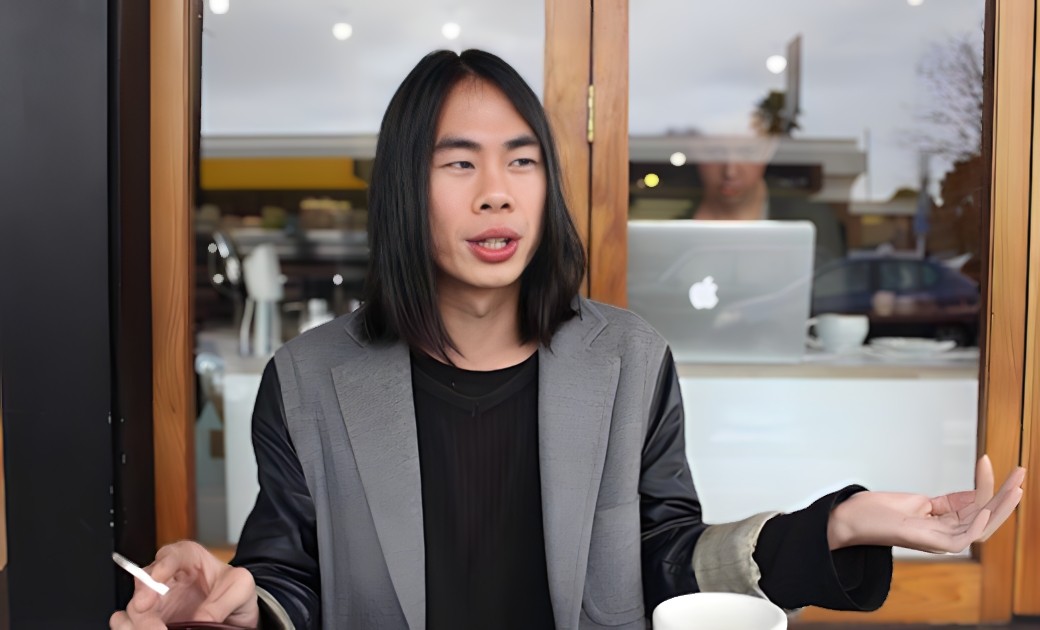 Man with dark shoulder length hair sitting outside a cafe gesticulating with a cigarette in one hand. 