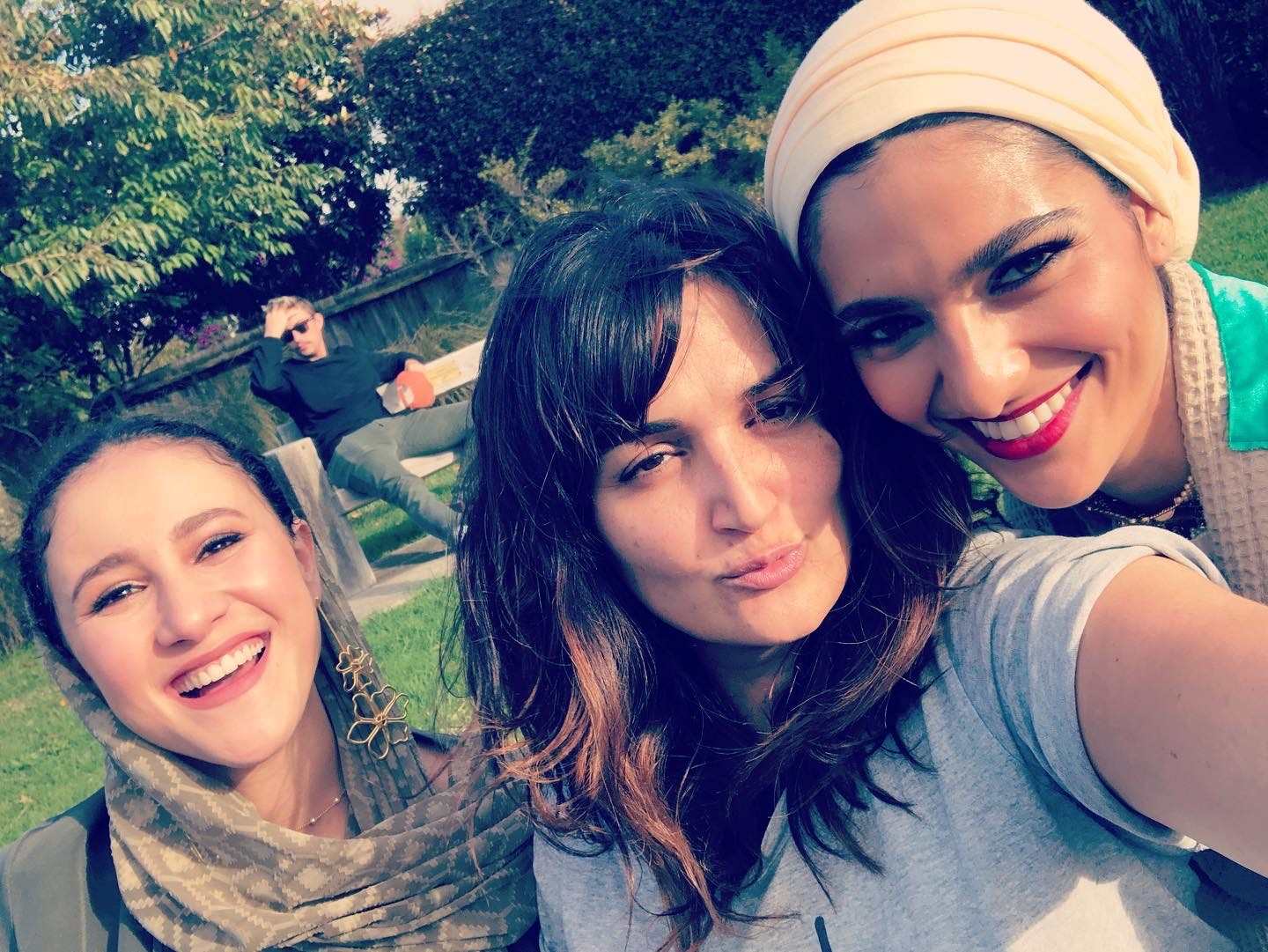 Three women smile and pose for a cheerful selfie in a sunny park, with a man lounging on a bench in the background.